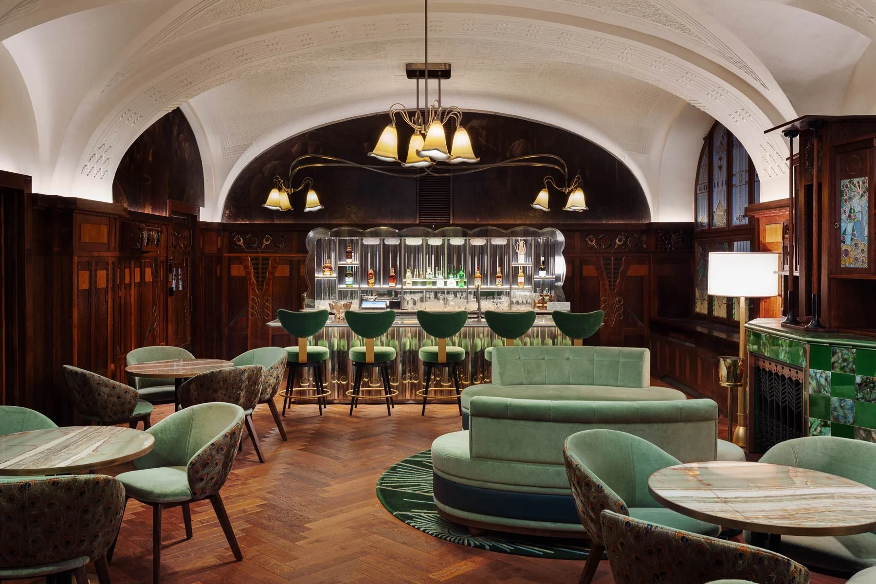 Elegantní bar v hotelu WPrague s tmavým dřevěným obložením, zelenými sametovými křesílky a barovým pultem s mramorovou deskou.