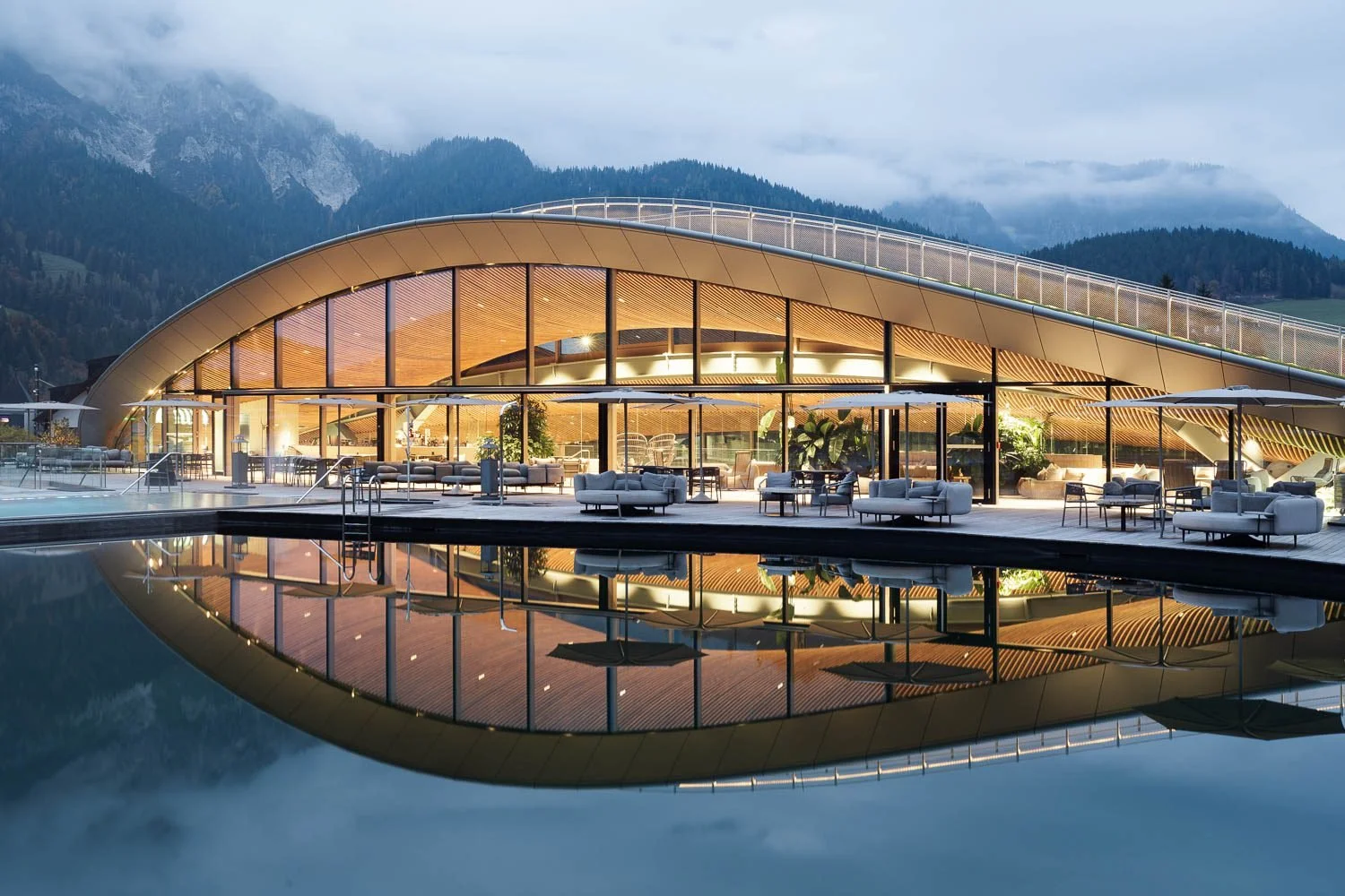 Architektonická fotografie wellness hotelu Krallerhof: večerní pohled na osvětlenou moderní budovu a venkovní koupací jezírko.