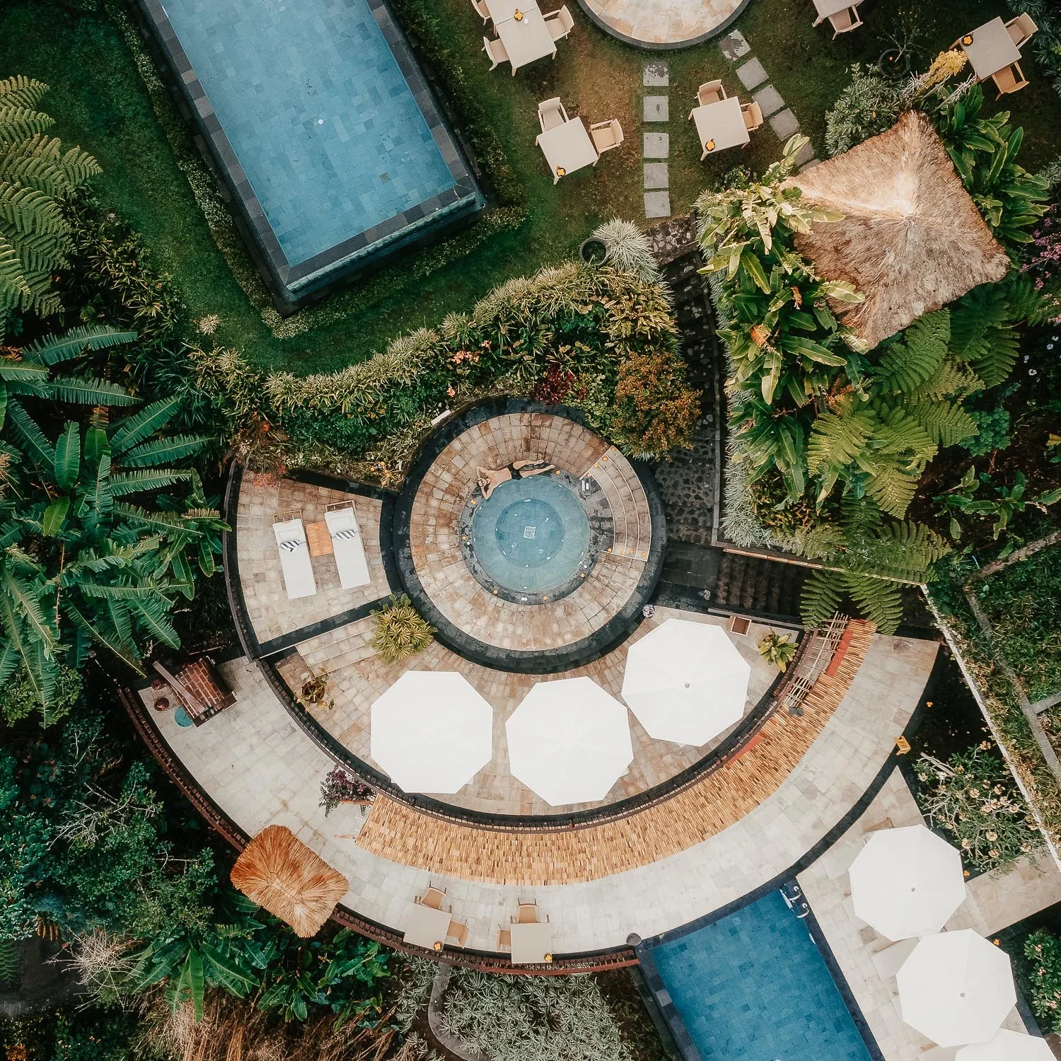 Letecký pohled z dronu na tropické letovisko s kruhovým bazénem, lehátky a palmami uprostřed svěží zeleně.