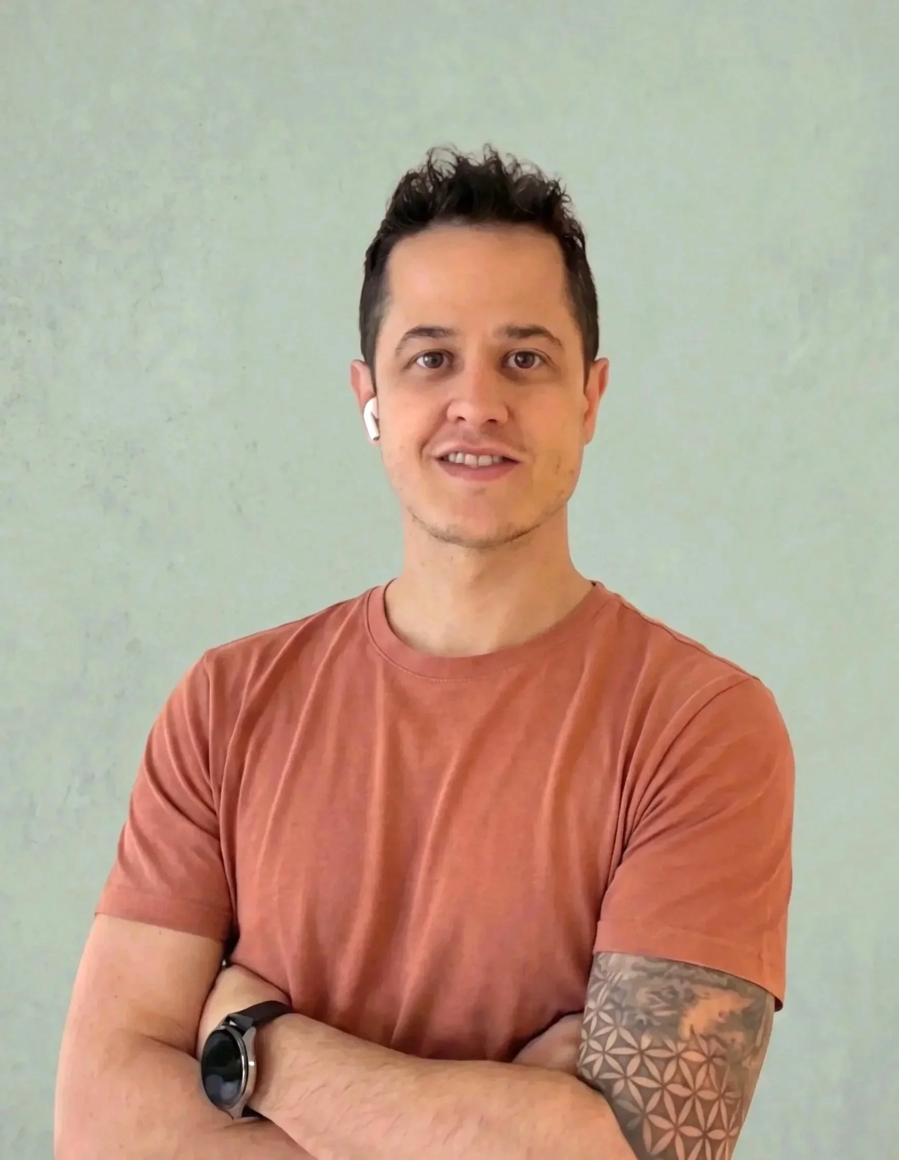 Joven hombre de cabello oscuro, usa audífonos blancos, camiseta naranja, reloj negro, con tatuaje en el brazo izquierdo, de fondo liso y sonrisa confiada.