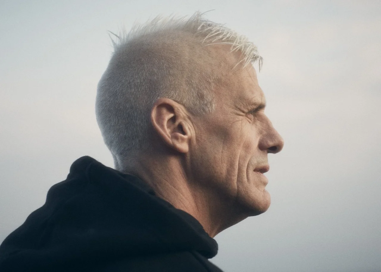 A middle-aged man with short, blond hair looking into the distance against a cloudy sky.