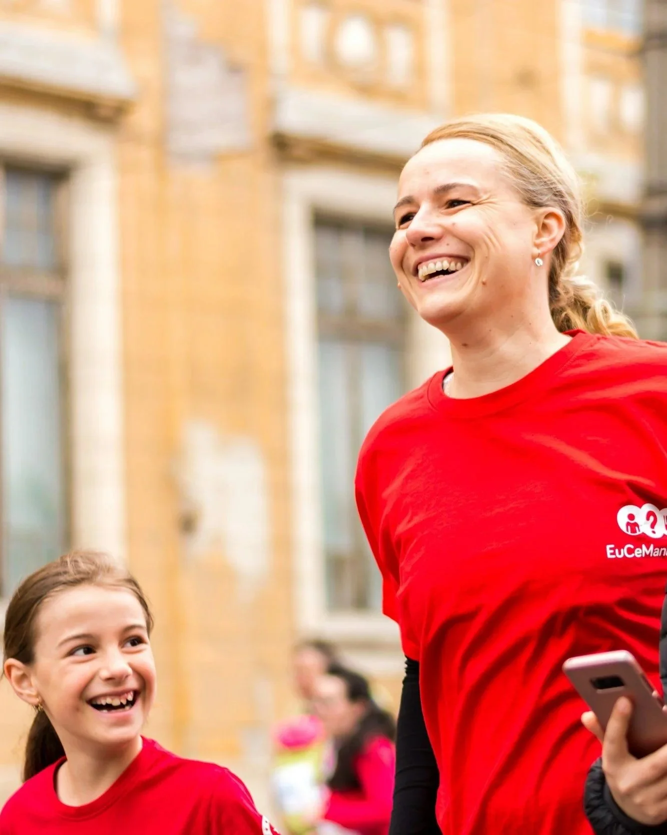 Smiling woman and girl in red shirts outdoors, likely at a community or sporting event, with the woman holding a smartphone.