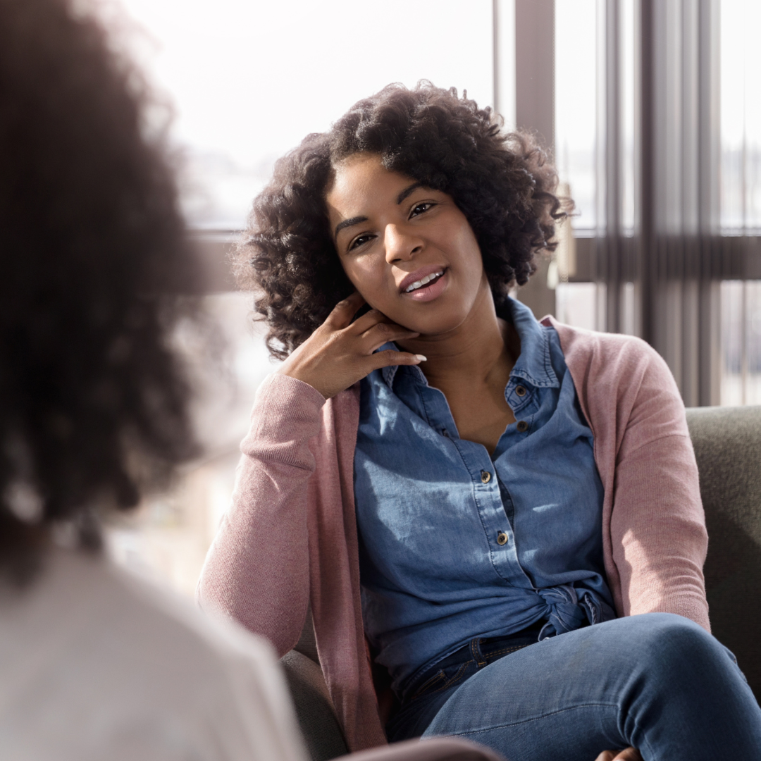 A woman with curly hair sitting on a sofa, talking to another woman in a bright room with large windows.
