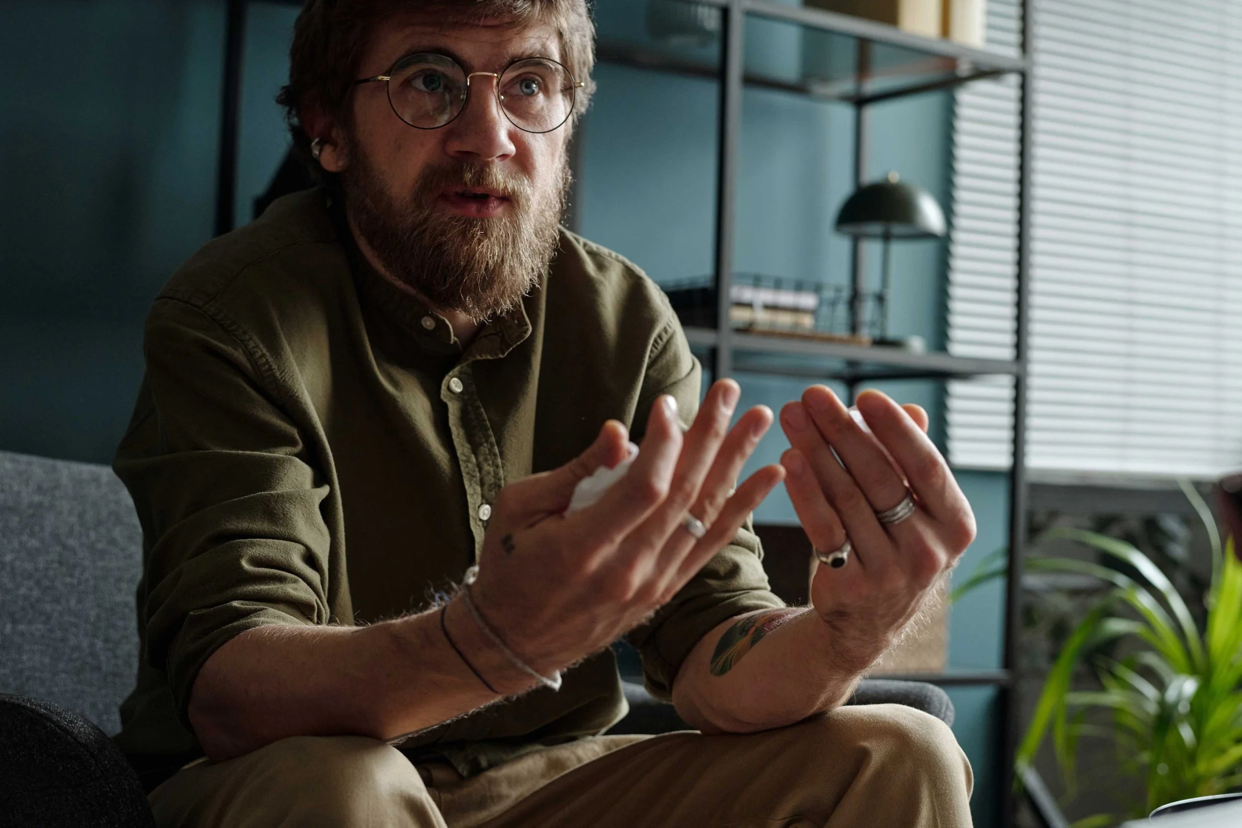 A man with glasses and a beard, wearing a green button-up shirt, sitting and gesturing with his hands in a modern room with shelves and plants.