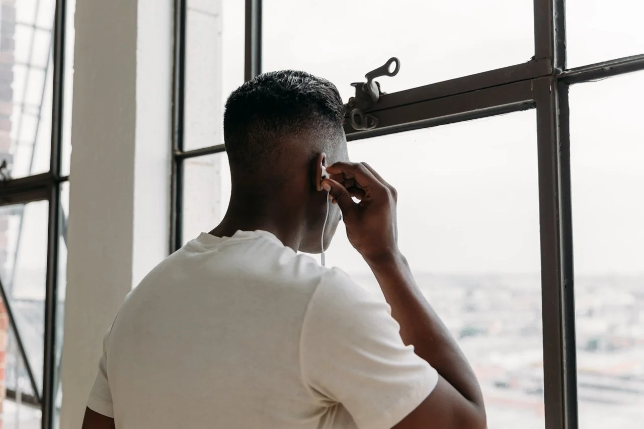 A young man with short hair and a white t-shirt holding a vintage phone to his ear, standing by a large industrial-style window overlooking a cityscape.