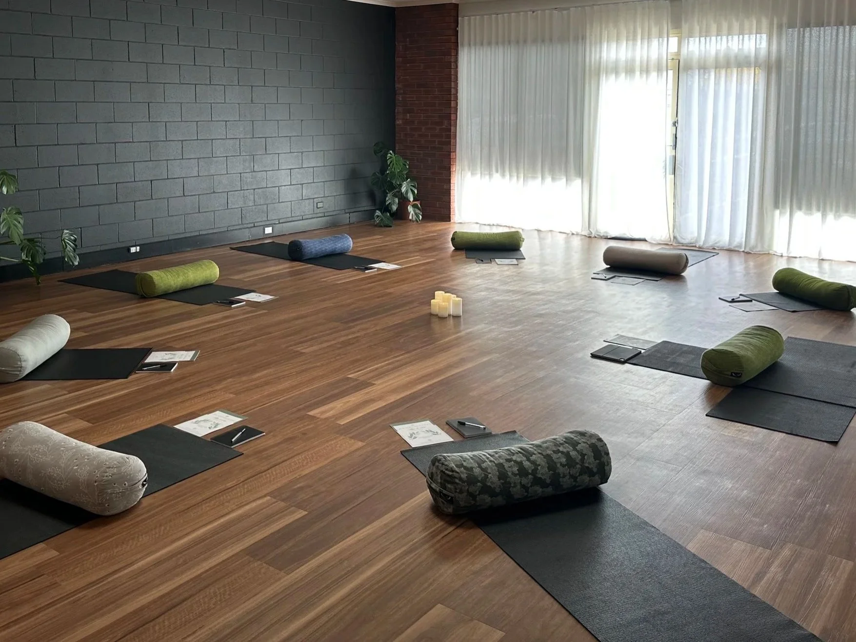 A spacious yoga studio with wooden floors, black yoga mats, and rolled-up cushions in various colors positioned at each mat. There are candles in the center and natural light streaming in through large curtains.