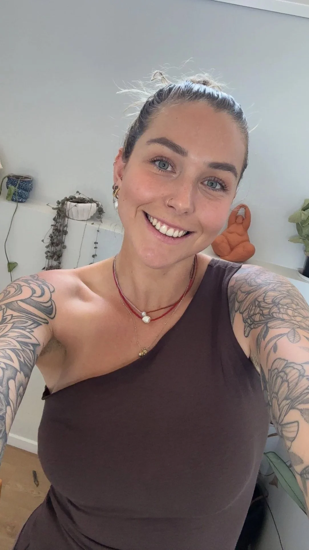 A woman taking a selfie with a big smile, wearing a brown tank top and layered necklaces, with tattoos on both arms and hoop earrings. Behind her are decorative plants and a small sculpture on a table.