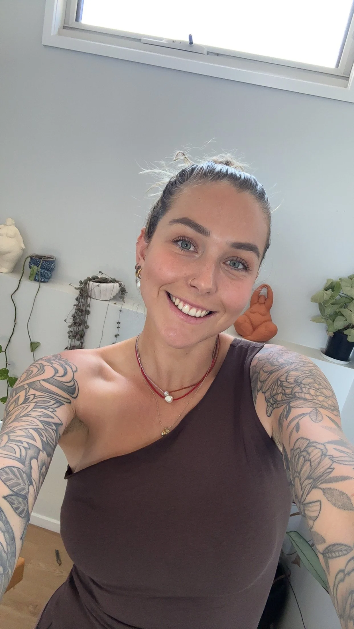 A woman with light skin, blue eyes, and dark hair pulled up, smiling and taking a selfie in a room with white walls and a window. She has tattoos on her arms, wears a sleeveless dark top, and has layered necklaces.
