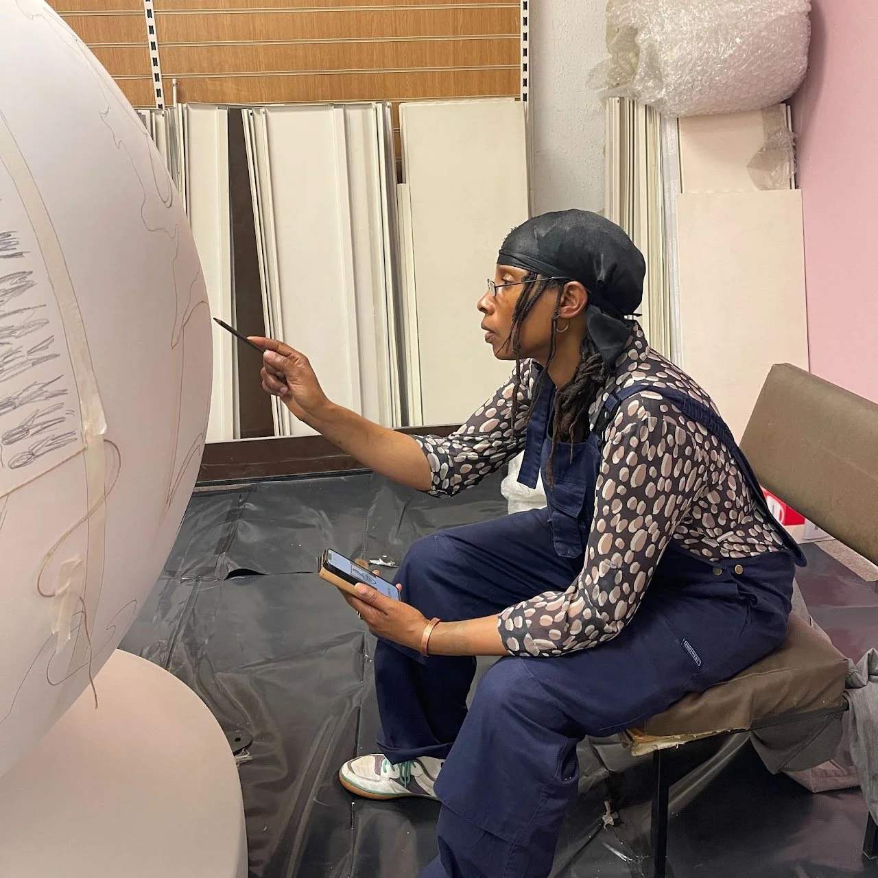 A woman with long dreadlocks, wearing glasses, a black head covering, and a patterned shirt, seated on a brown chair, pointing at an artwork with a pencil while holding a smartphone in her other hand, in an indoor studio or classroom setting.