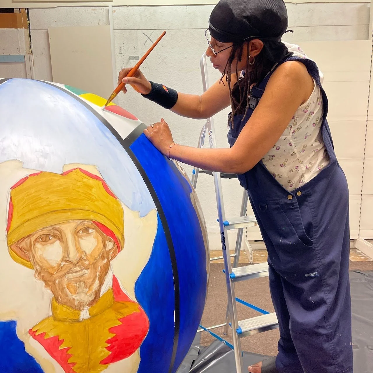 An artist painting a large globe with a portrait of a man wearing a yellow and red turban.