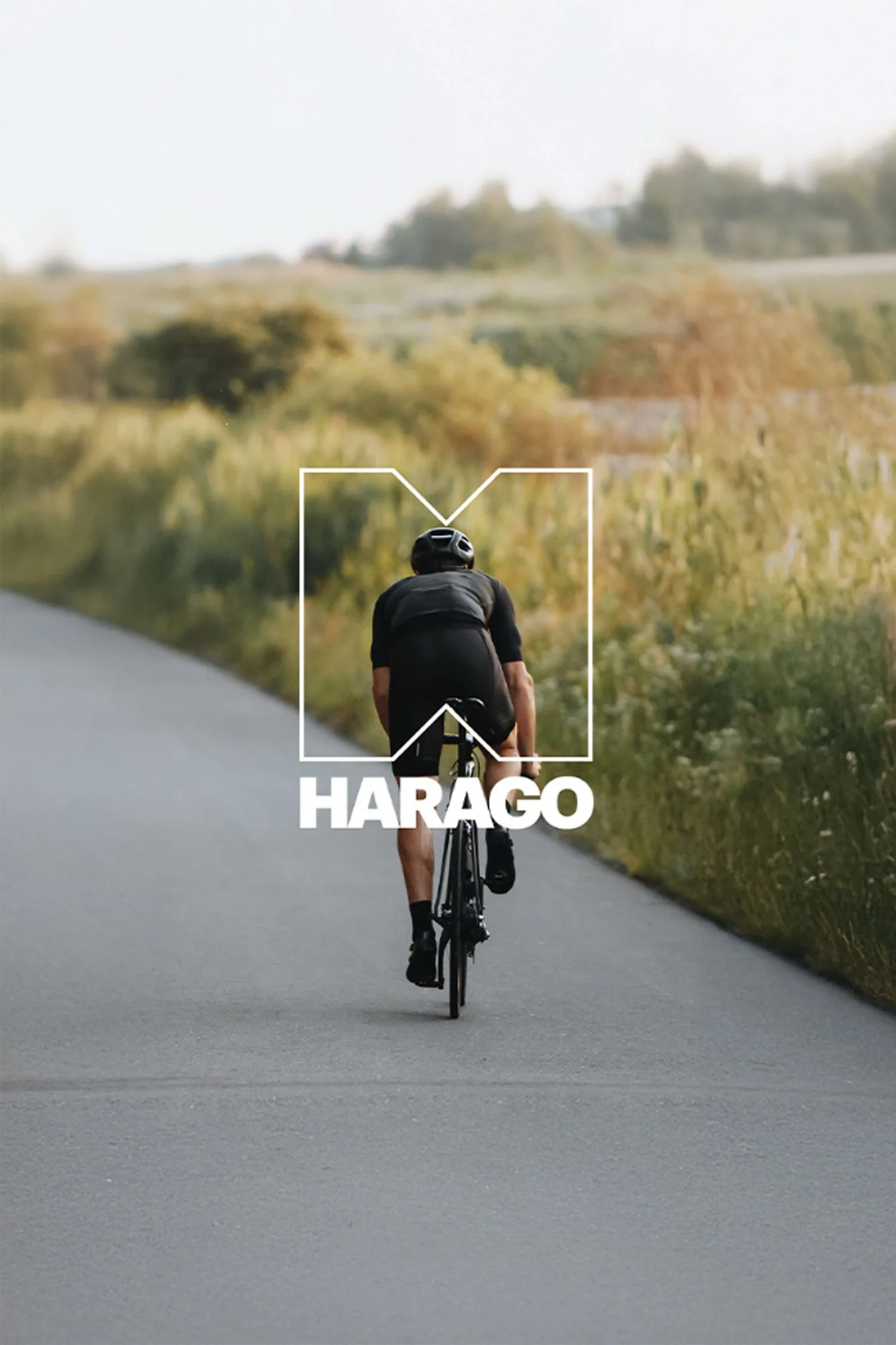 Photo d'un  cycliste vu de dos, roulant sur une route dans un paysage rural avec des champs et des arbres, avec le texte 'HARAGO' en gros caractères blancs à l'avant-plan.