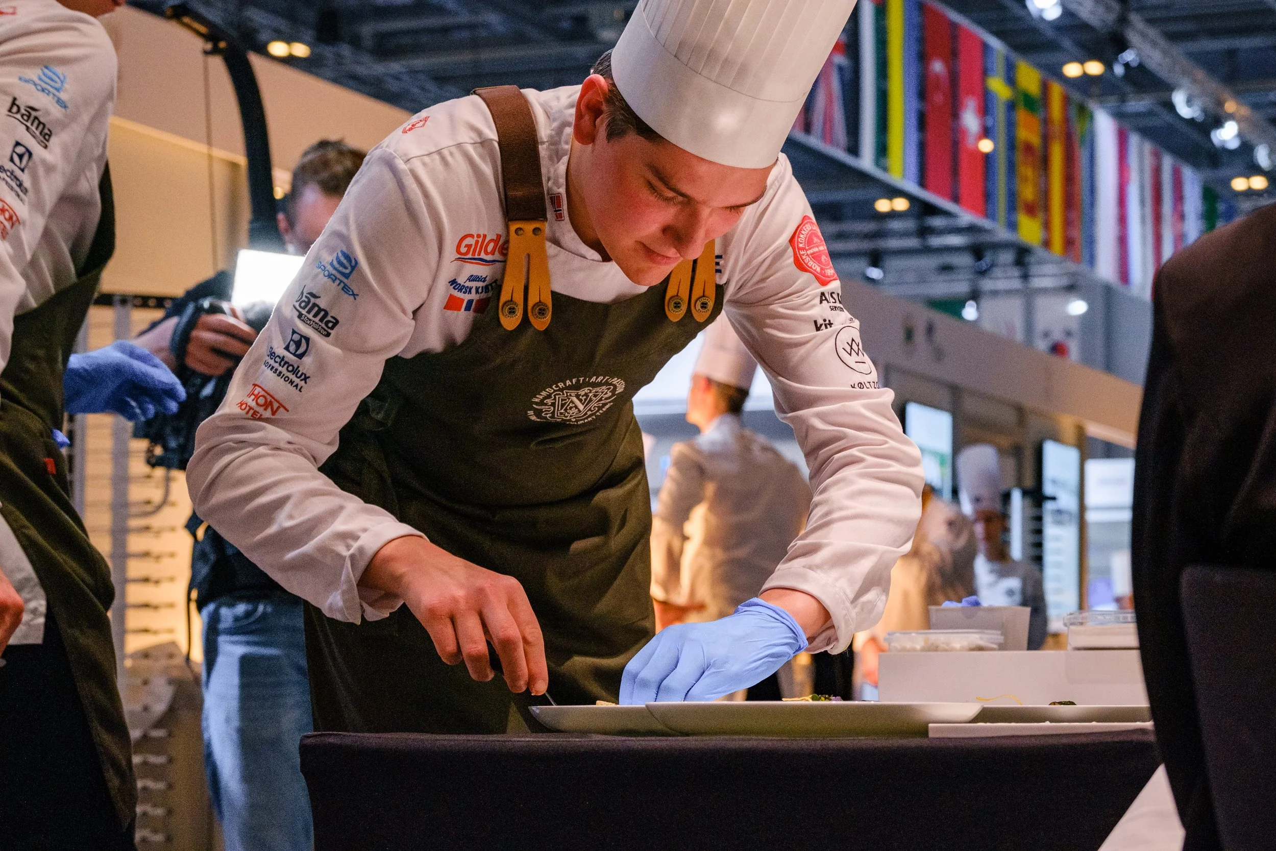 Kokk Andreas Fjellheim i hvit uniform og kokkehatt forbereder mat under IKA Culinary Olympics, med flere folk og nasjonalflagg i bakgrunnen.
