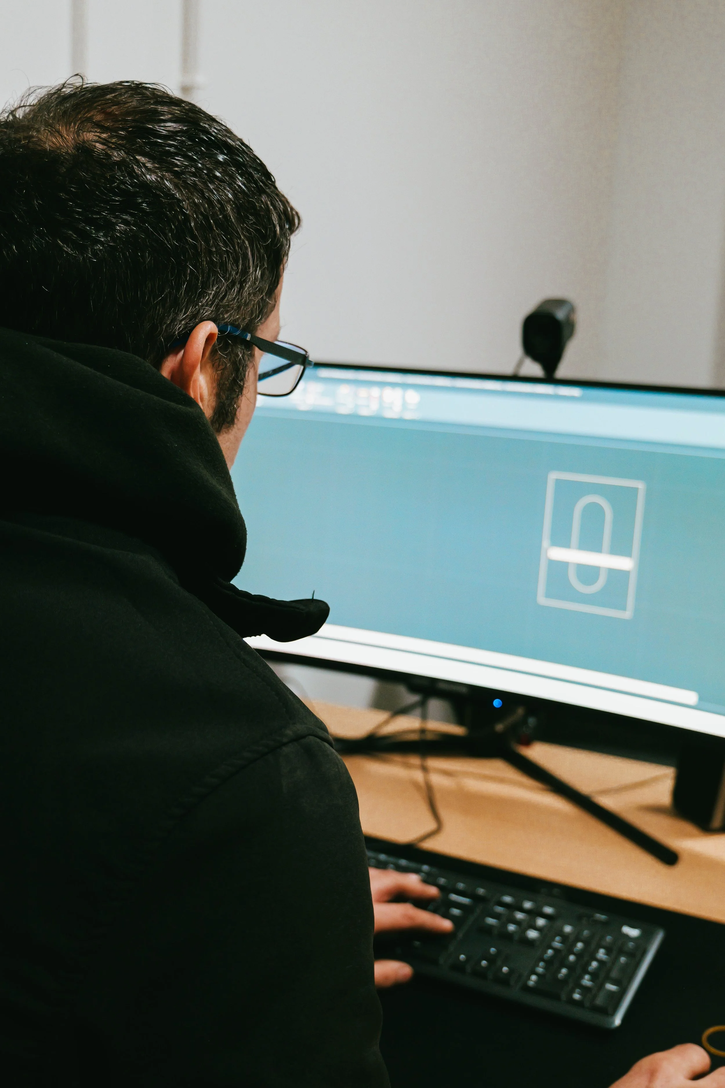 A person wearing glasses and a black hoodie sitting at a desk using a computer with a large monitor to play a video game.