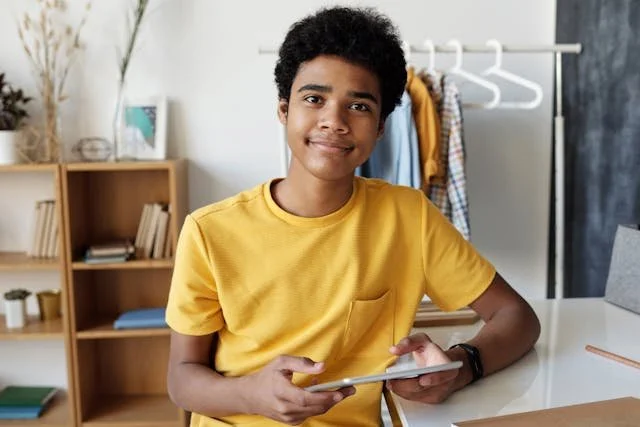 Teenage boy in a yellow tshirt is holding an ipad and smiles into the camera