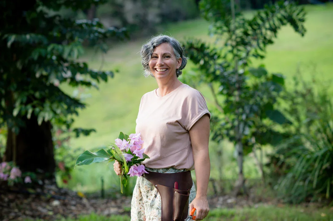 A smiling woman in casual clothes holding a bouquet of pink flowers in a lush green garden.