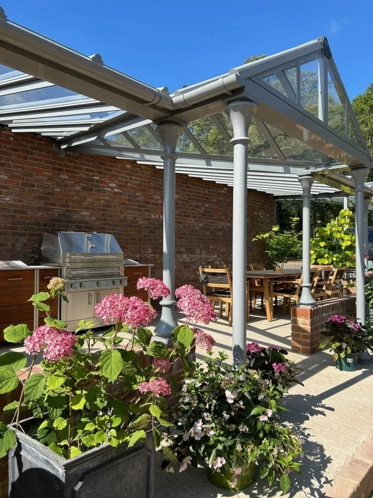 An outdoor patio area with a clear glass roof supported by white columns, a brick wall in the background, a stainless steel grill, a wooden dining table with chairs, and potted flowering plants in the foreground.