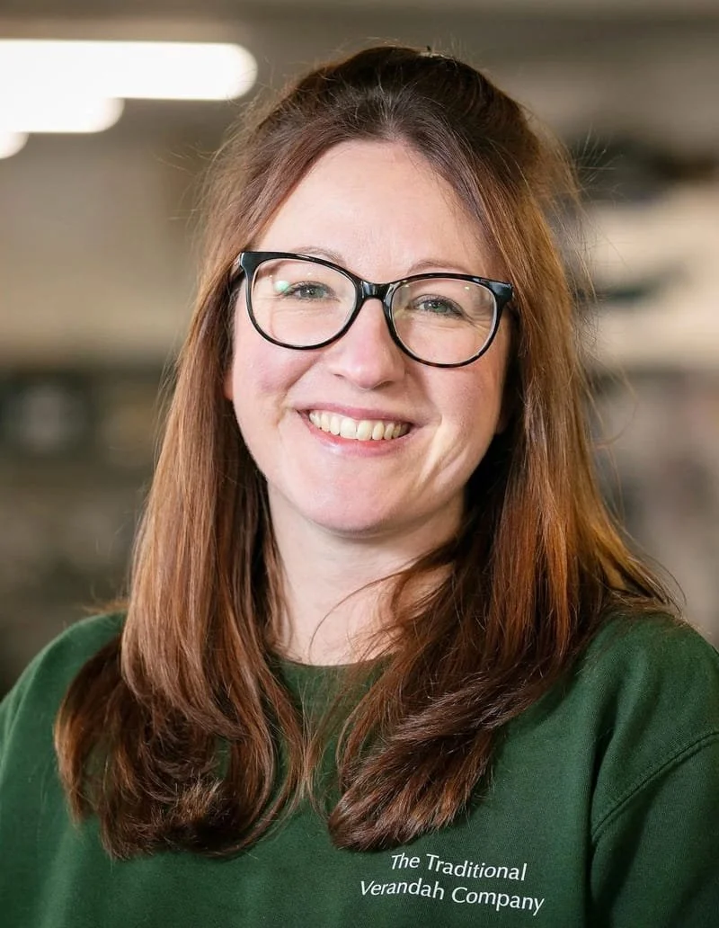 A woman with long reddish-brown hair, wearing glasses and a green shirt that reads 'The Traditional Verandah Company,' smiling at the camera in an indoor setting.