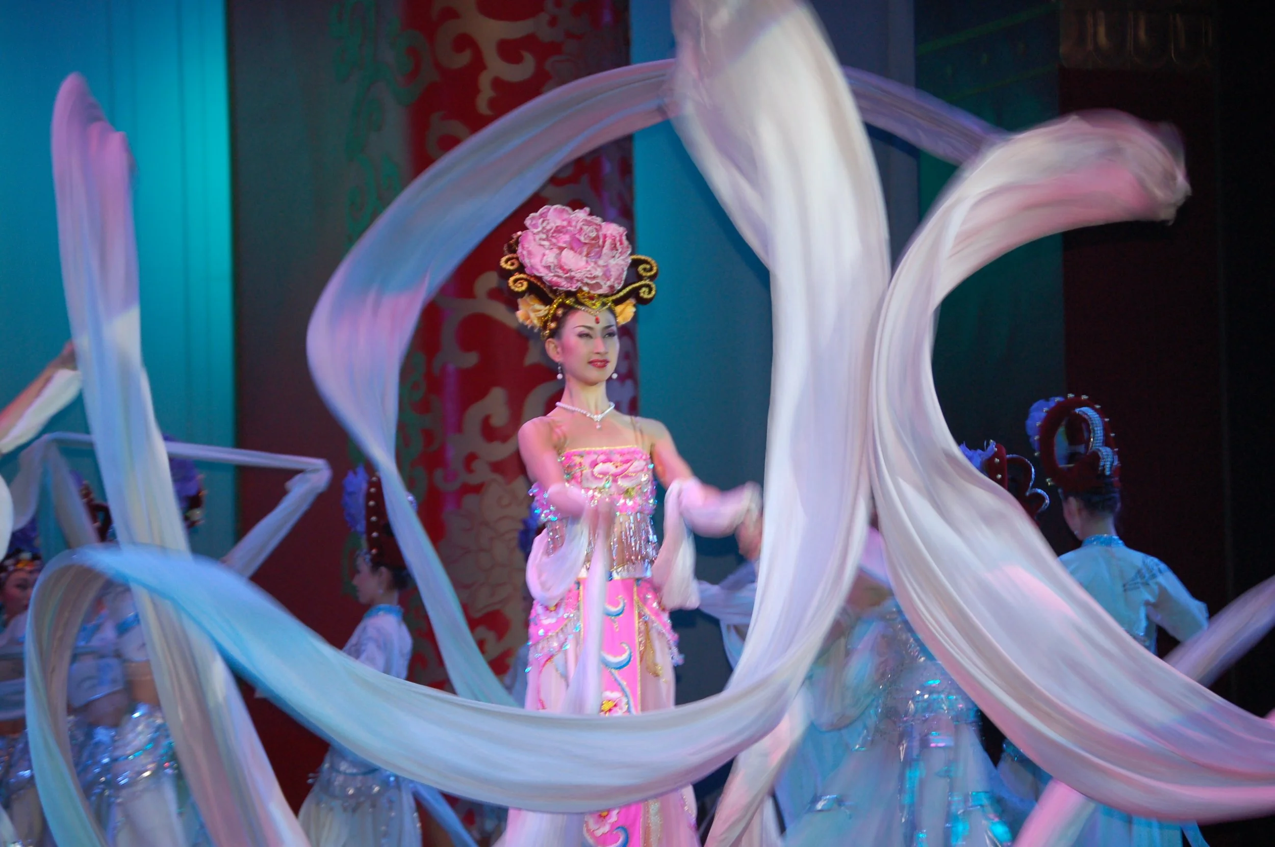 Performer dressed in traditional Asian costume with elaborate headdress, dancing with flowing white silk ribbons on stage during cultural performance.