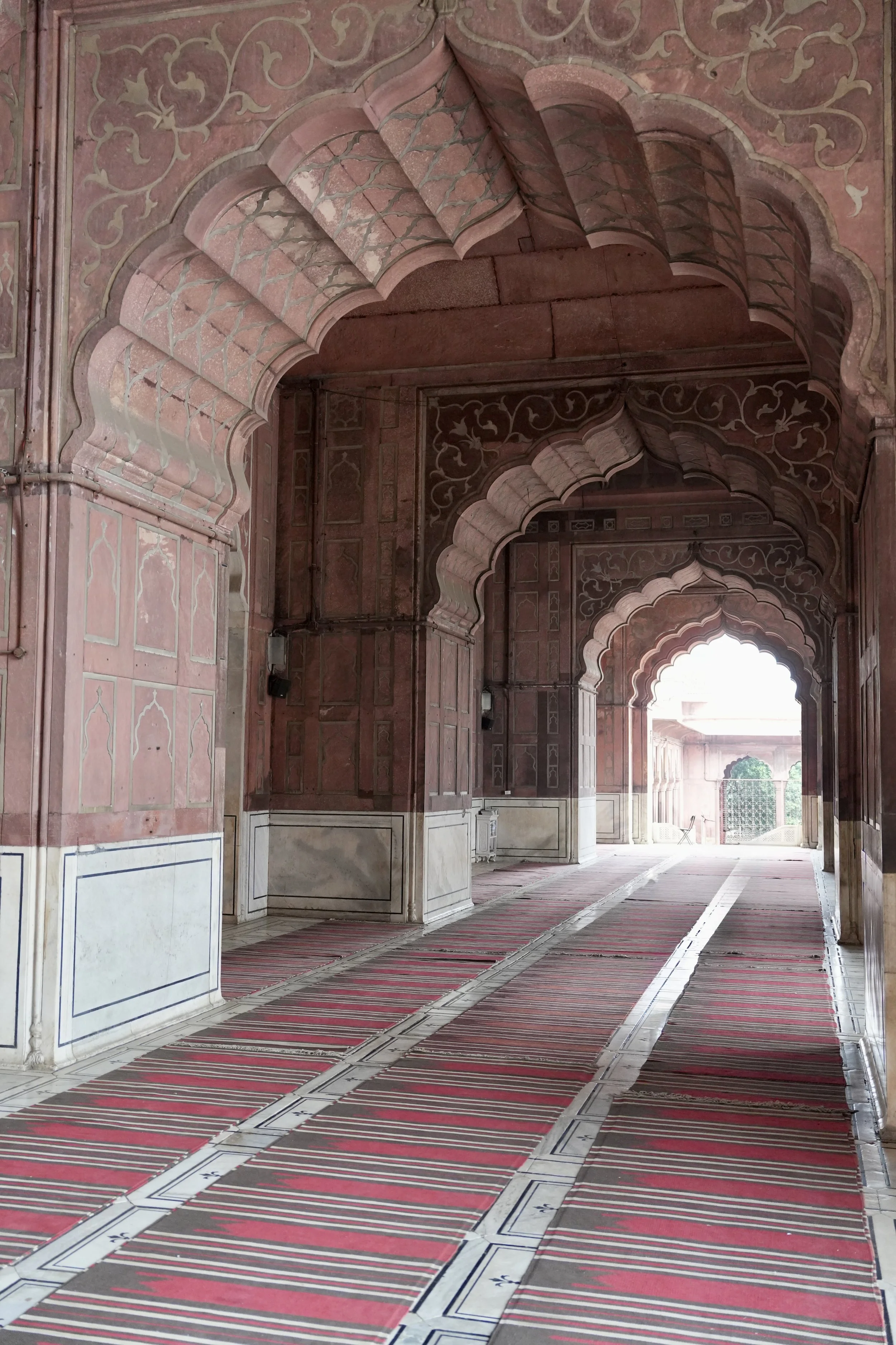  Ornate red sandstone archways inside a mosque, with intricate carved patterns on the walls and ceiling, leading down a corridor lined with red striped prayer carpets towards a bright courtyard beyond. 