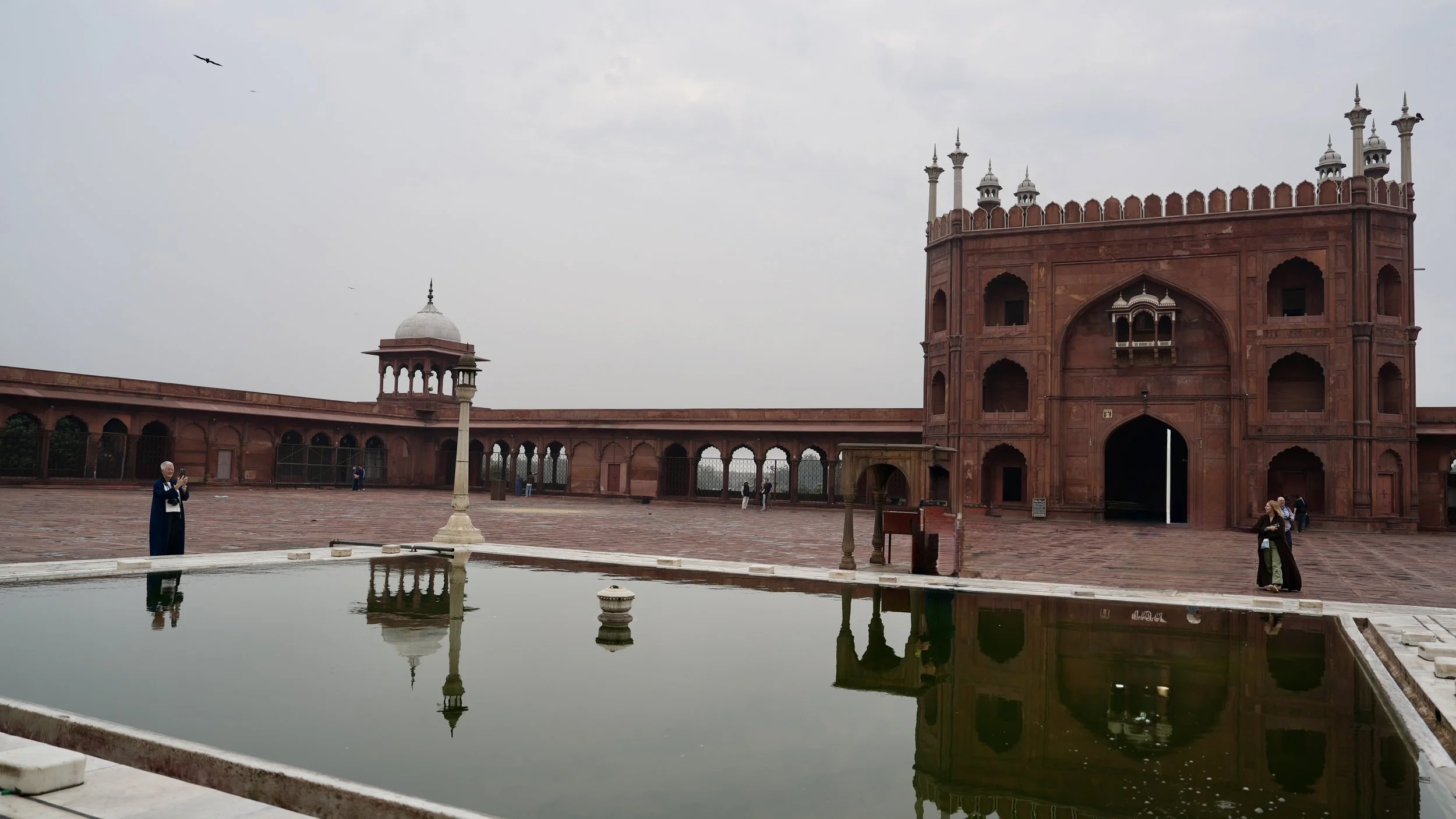  Wide courtyard of a red sandstone mosque with arched colonnades and a domed pavilion, seen across a rectangular ablution pool reflecting the buildings, with a few visitors standing quietly around the edge. 