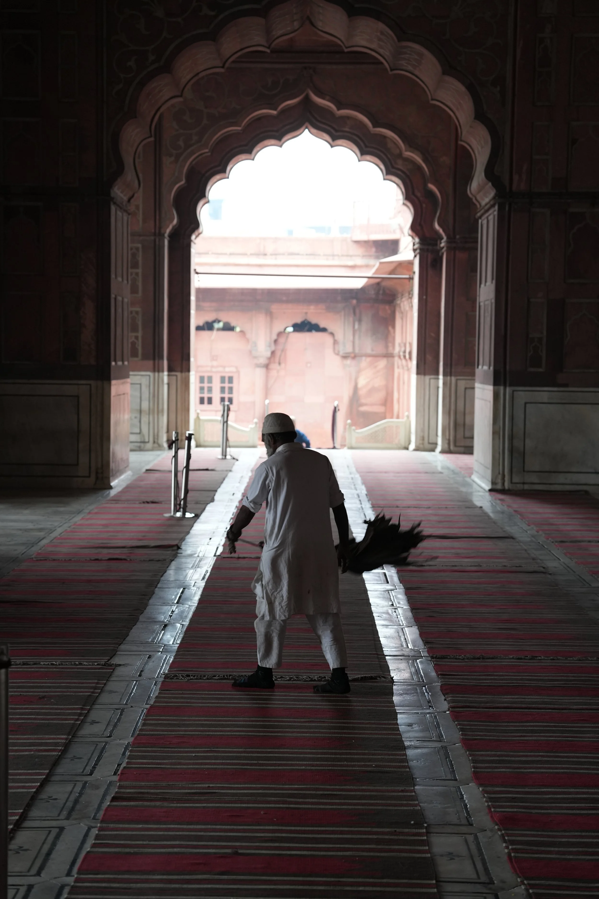  Silhouette of a man in white clothing sweeping the carpet inside a mosque, standing beneath an ornate arched doorway, with patterned red prayer mats on the floor and soft light coming through the courtyard beyond. 