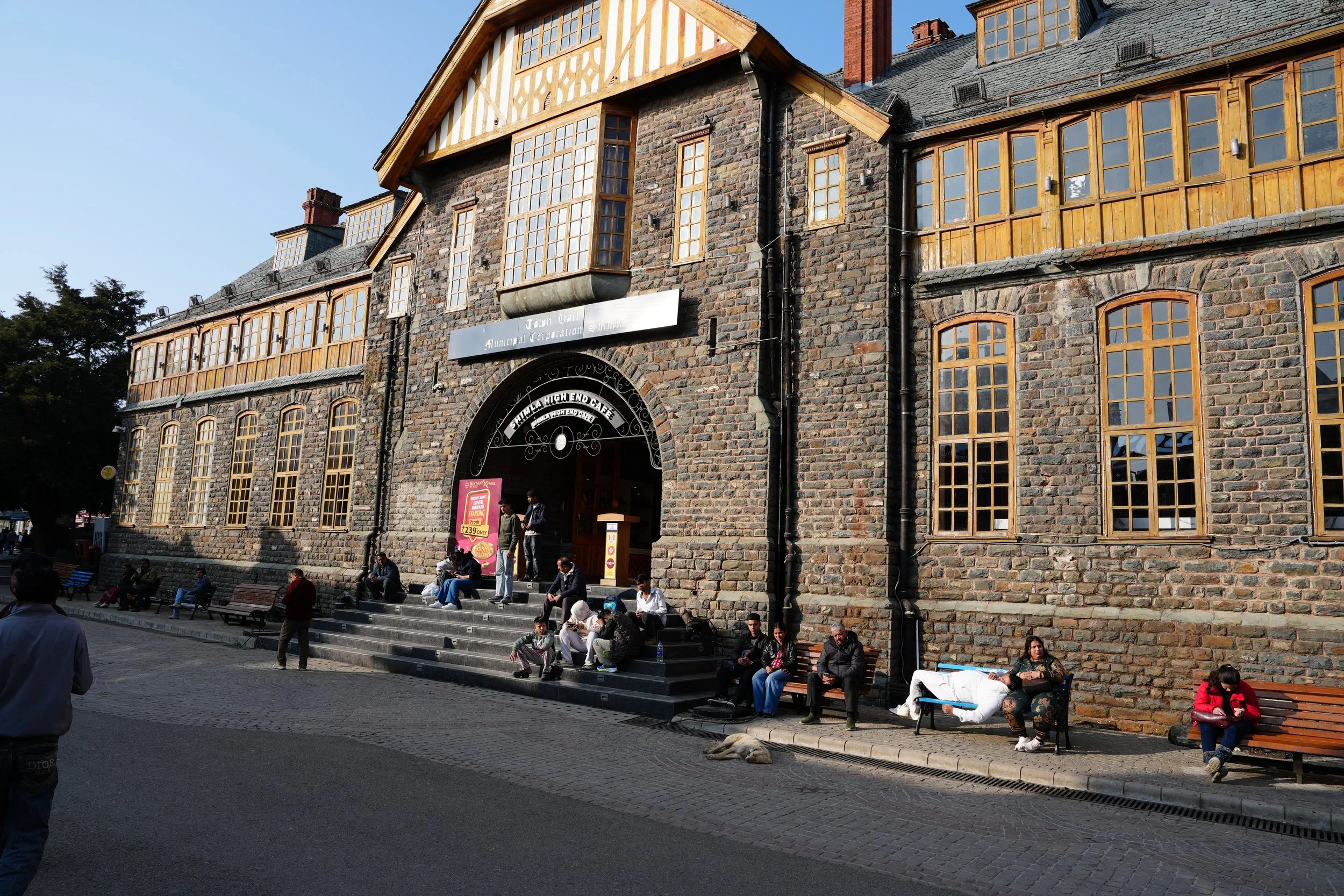  Historic stone building on the Mall in Shimla with large wooden-framed windows and an arched entrance, with people sitting on benches and steps in the afternoon sun outside the Indian Coffee House. 
