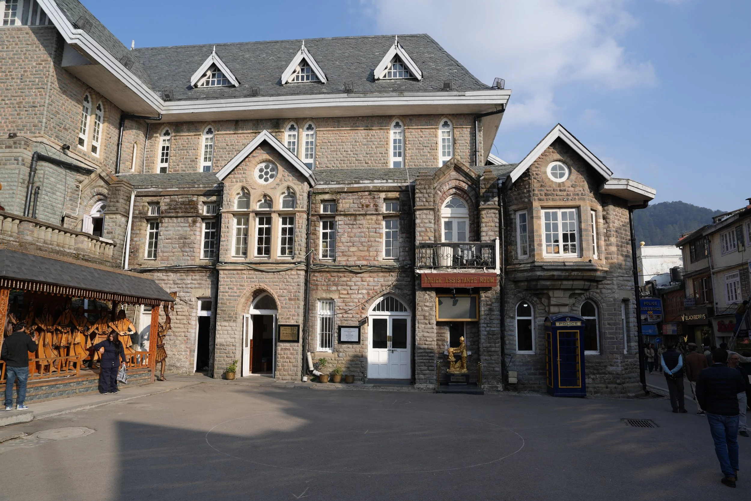  Colonial-era stone building on the Ridge in Shimla with arched windows and steep rooflines, with a small square in front and people walking past shops and a police assistance booth.   