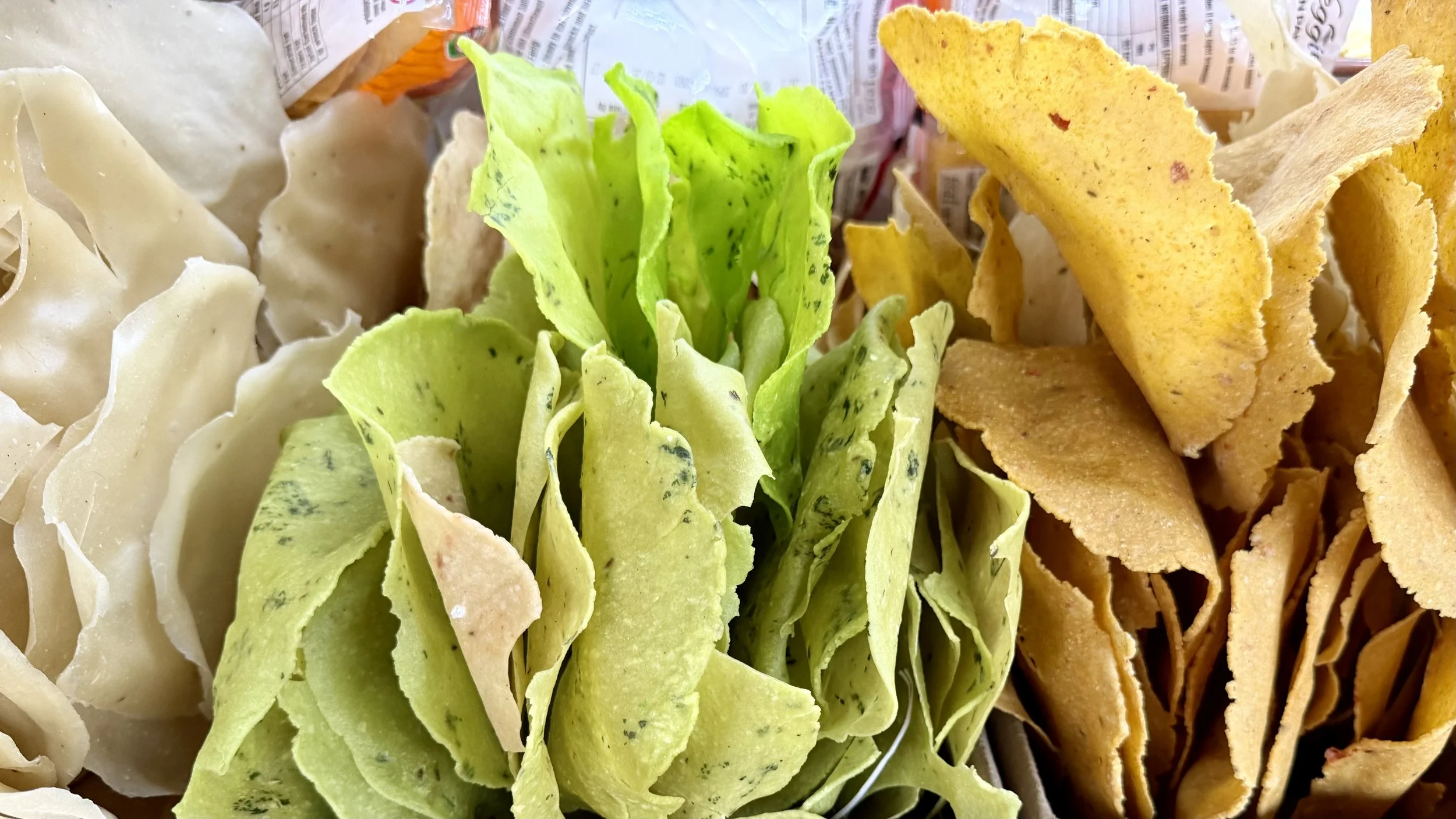 Close-up of crisp snacks in pale green, golden and off-white colours piled in bags at a market stall.