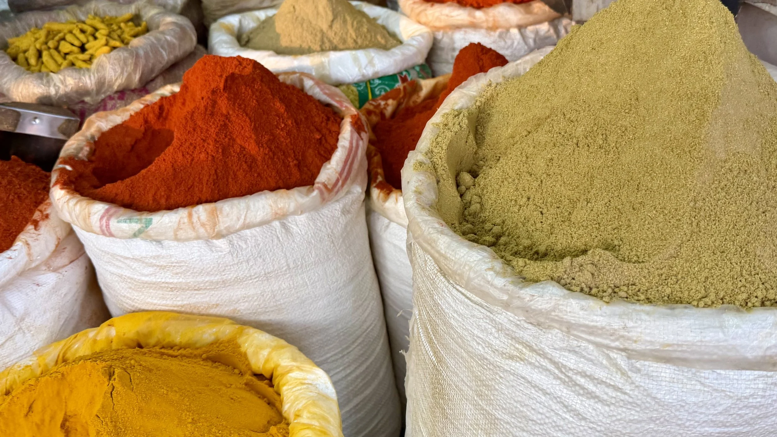 Large white sacks filled with brightly coloured powdered spices stacked together in a market.