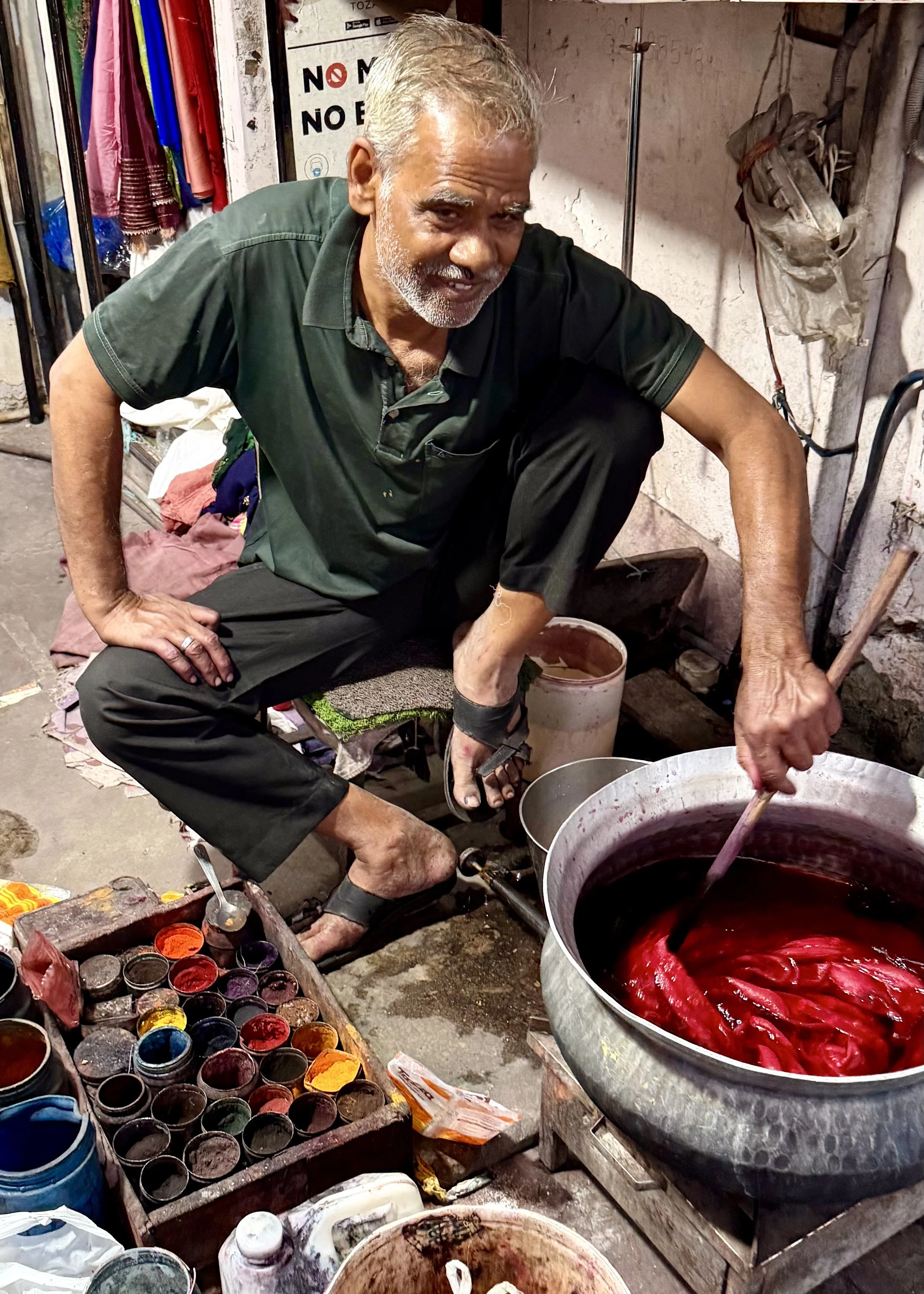 Man crouching beside a large metal pot of red dye, stirring fabric while small tins of pigment sit nearby.