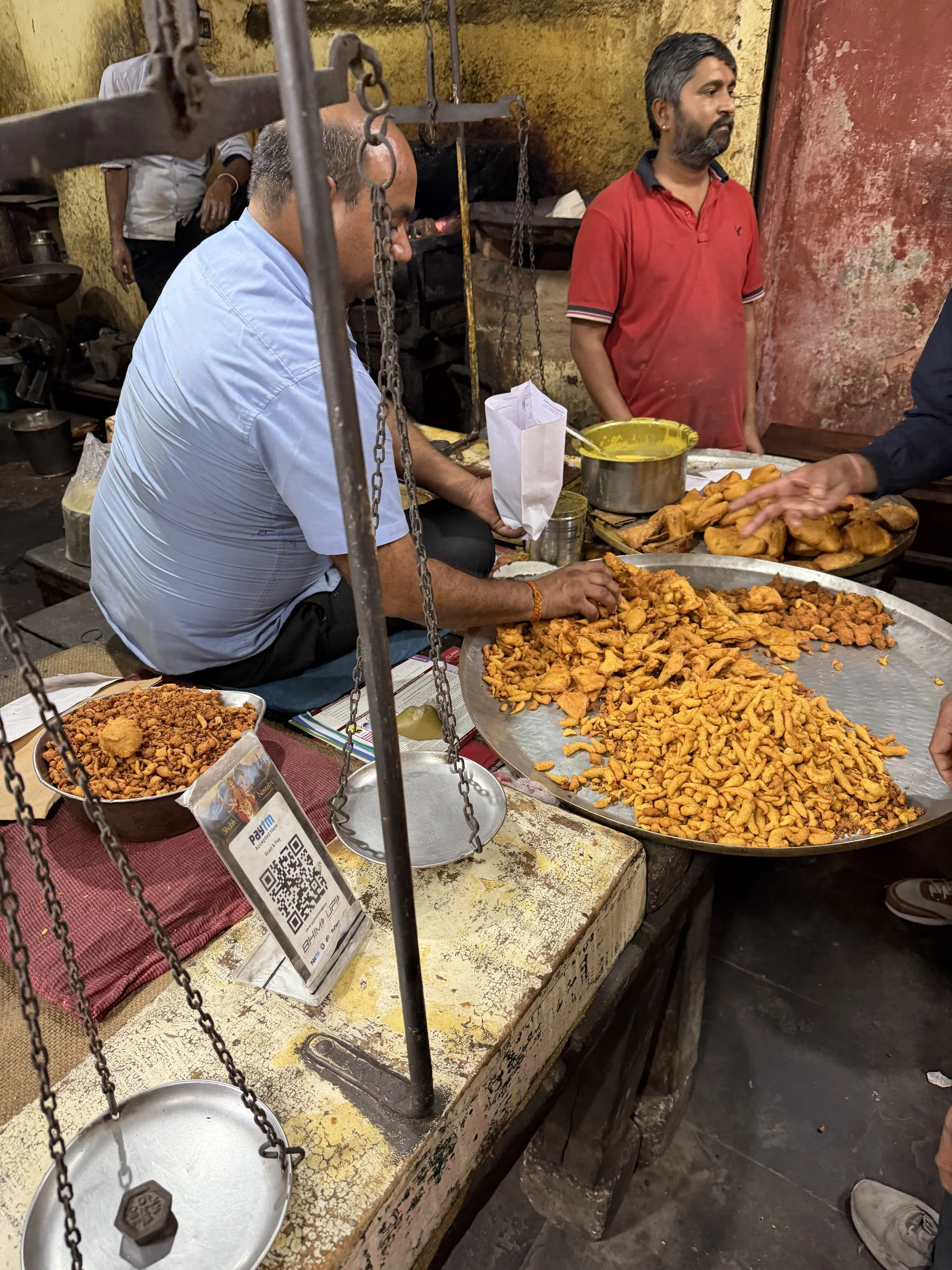 Snack seller working behind a worn counter with fried savoury snacks, weighing scales and a QR payment sign.