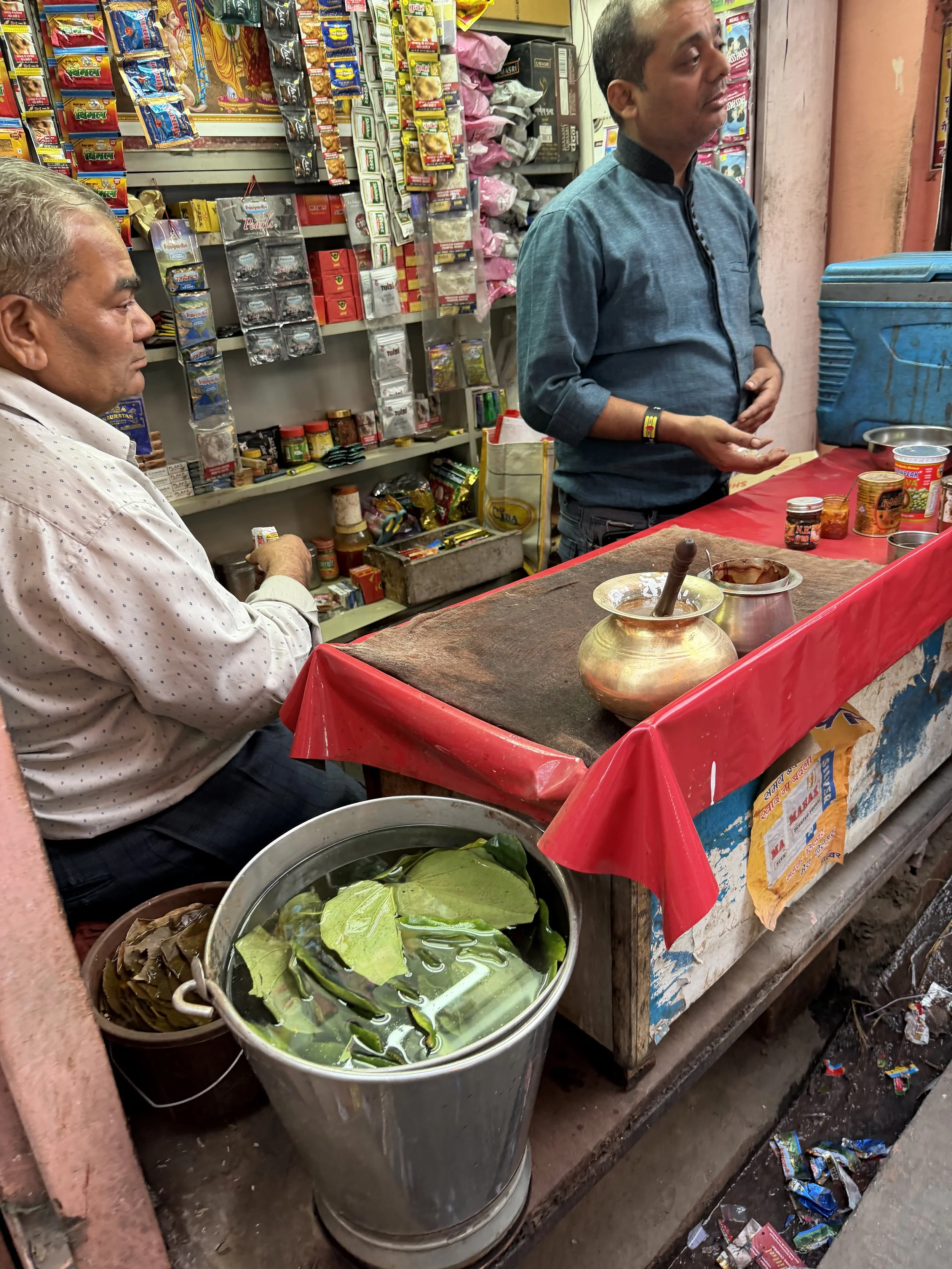 Vendor standing at a small paan stall with betel leaves soaking in a metal bucket and packets hanging behind the counter.