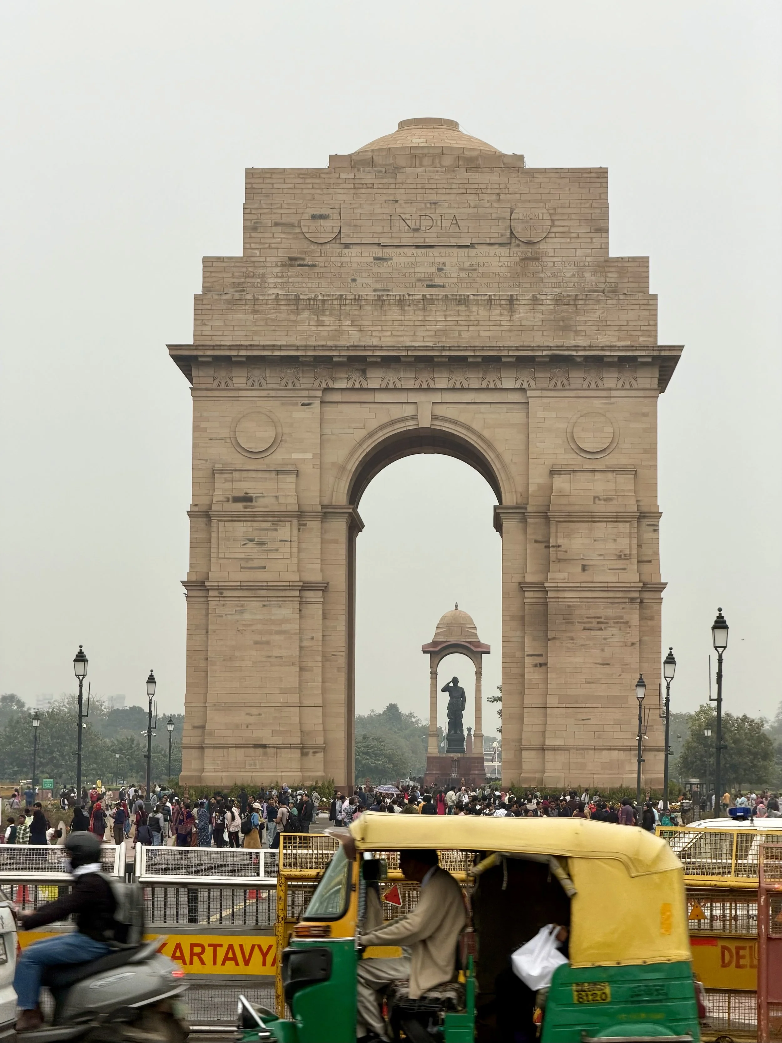 India Gate war memorial in Delhi seen through busy traffic, with yellow and green auto-rickshaws passing in the foreground and the arch framing a distant statue beneath the monument.