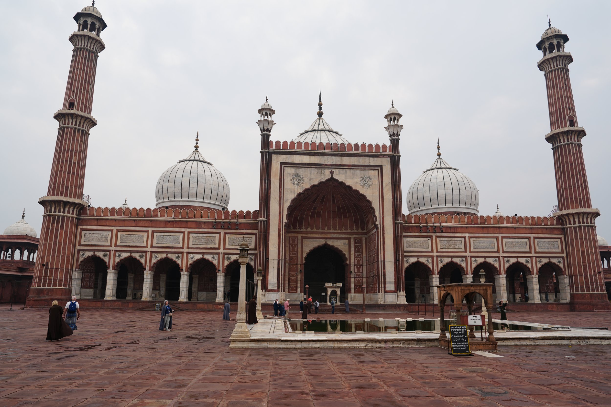 Jama Masjid, Old Delhi Front view of a large Mughal-style mosque with red sandstone walls, white domes, and two tall minarets, seen across a wide courtyard with a small ablution pool in the foreground and a few visitors walking around.