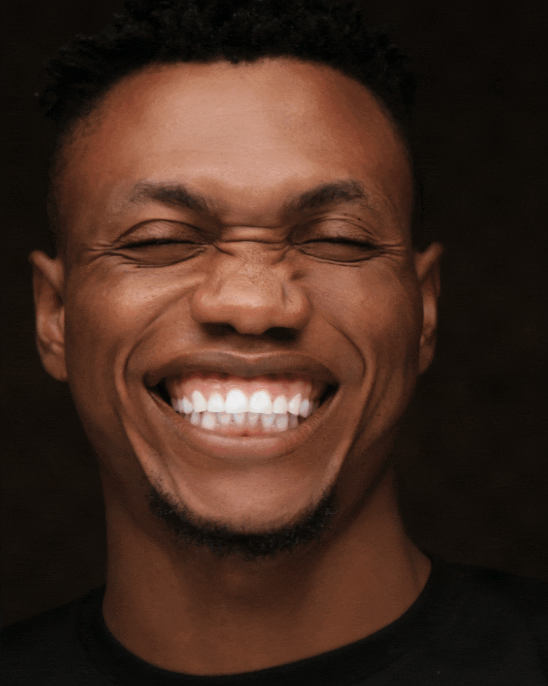 A young man with short, curly hair and a beard smiling broadly with eyes closed, showing his teeth.
