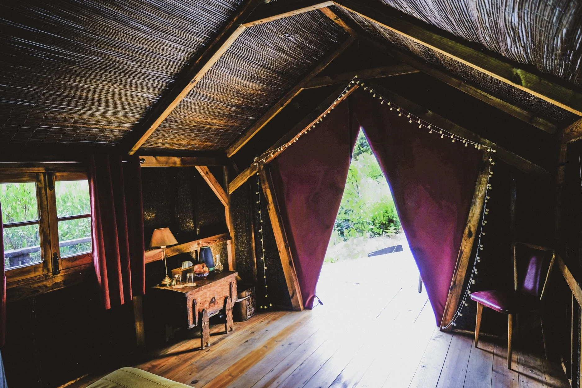 Interior of a cozy wooden cabin with a sloped ceiling, purple curtains framing an open doorway, a window with red curtains, and a small wooden table with a lamp and decorative items inside, with outside greenery visible through the doorway.