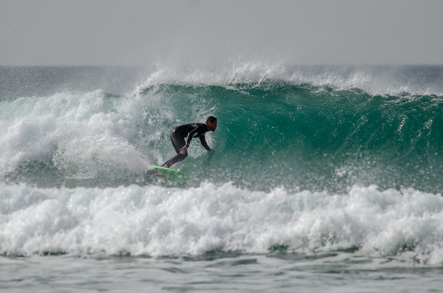 Tiago Madeira in a black wetsuit riding a large, greenish wave on a surfboard in the ocean.