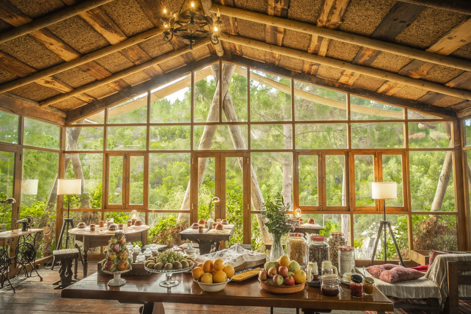 Interior of a rustic wooden room with large glass windows showing green trees outside, decorated with table setups and jars of food, and lit by a chandelier and standing lamps.