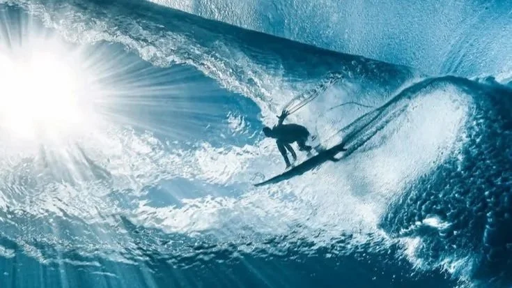 A person surfing on a large ocean wave with sunlight shining from the left.