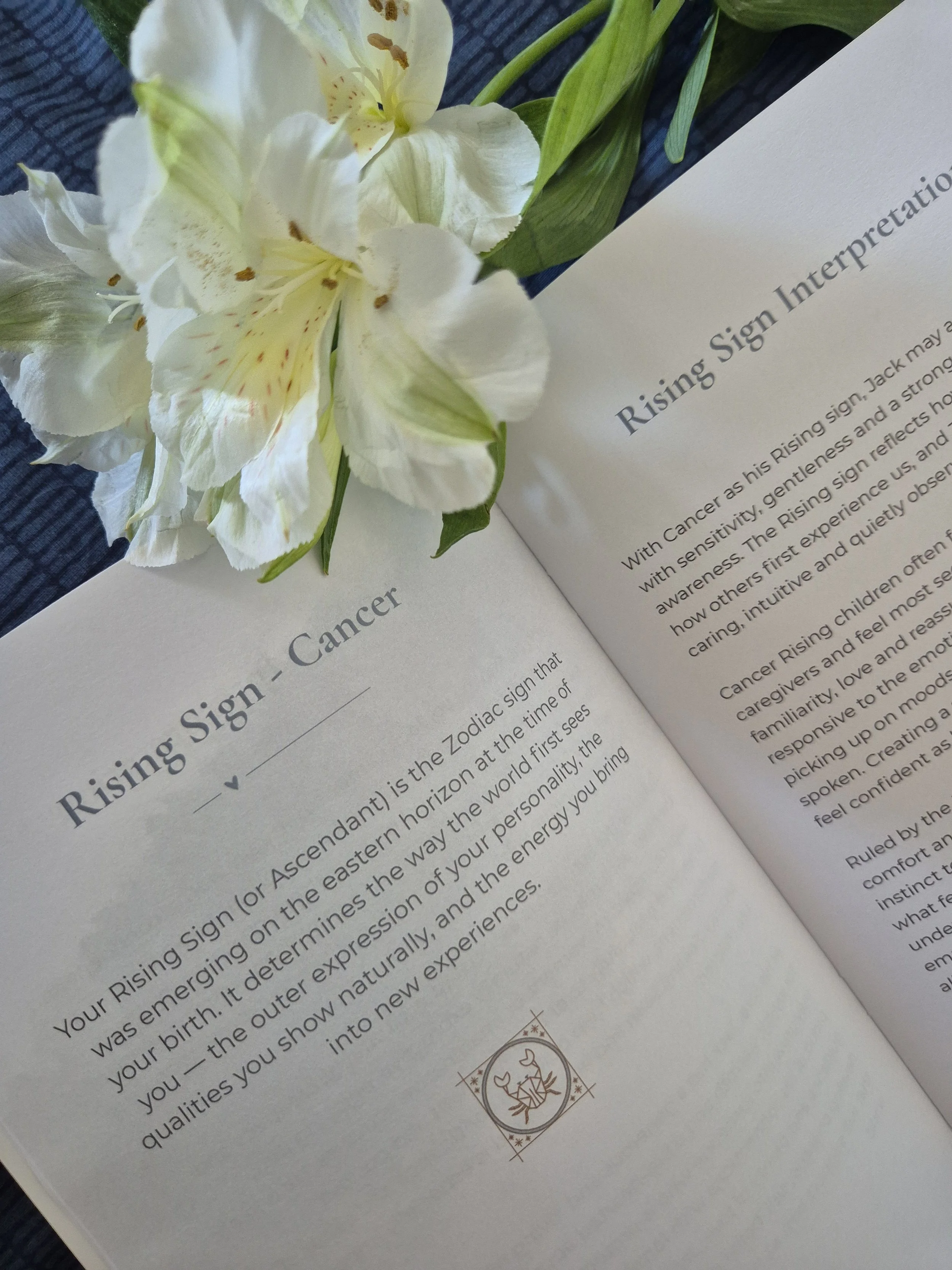 Open baby astrology book on a table with a white flower arrangement nearby, showing a page about the zodiac sign Cancer.