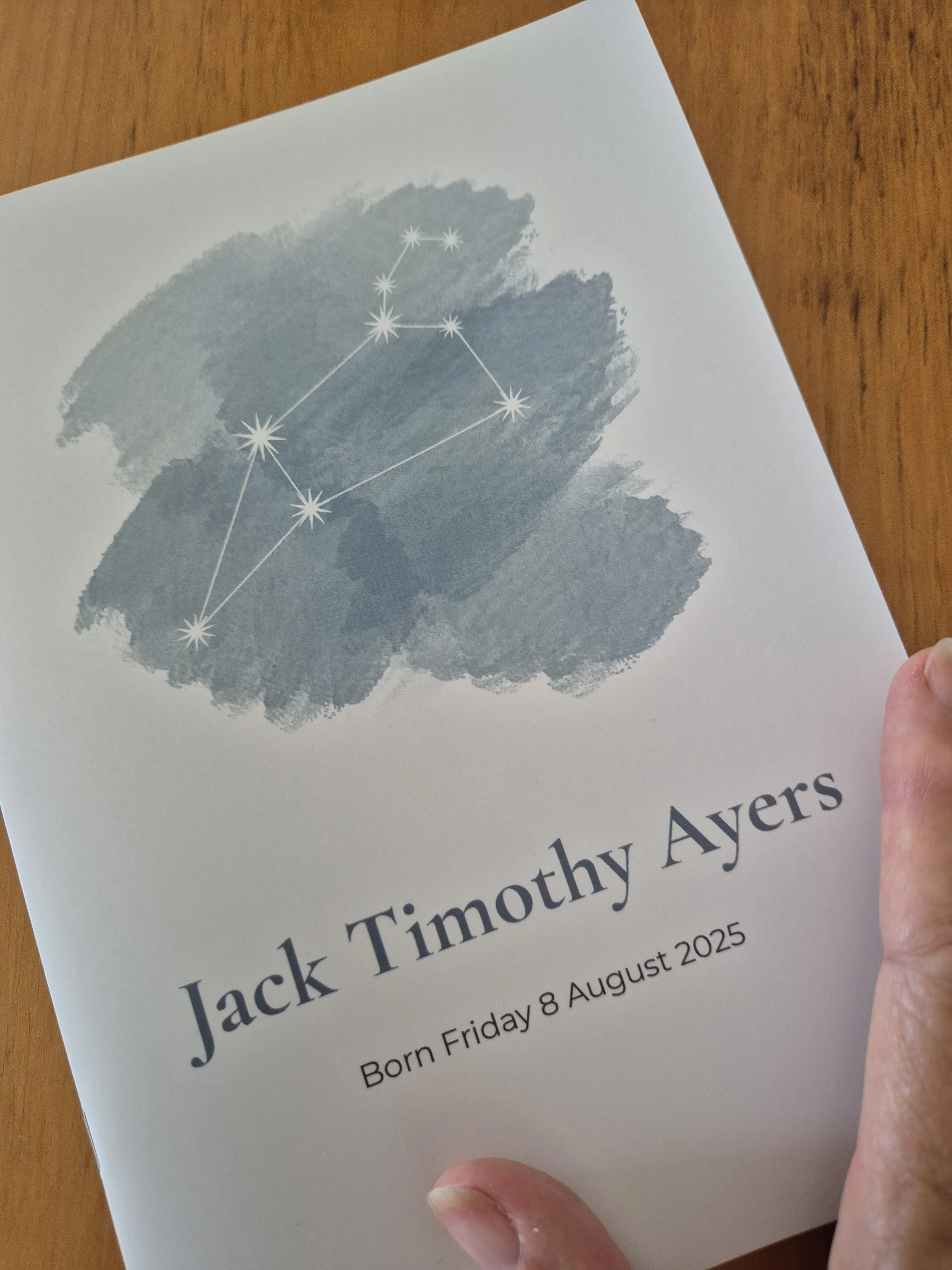 A printed card honoring Jack Timothy Ayers on his birthday, August 8, 2025. The card features a watercolor design with a star constellation illustration above his name and birth date, all held by a person's hand over a wooden surface.
