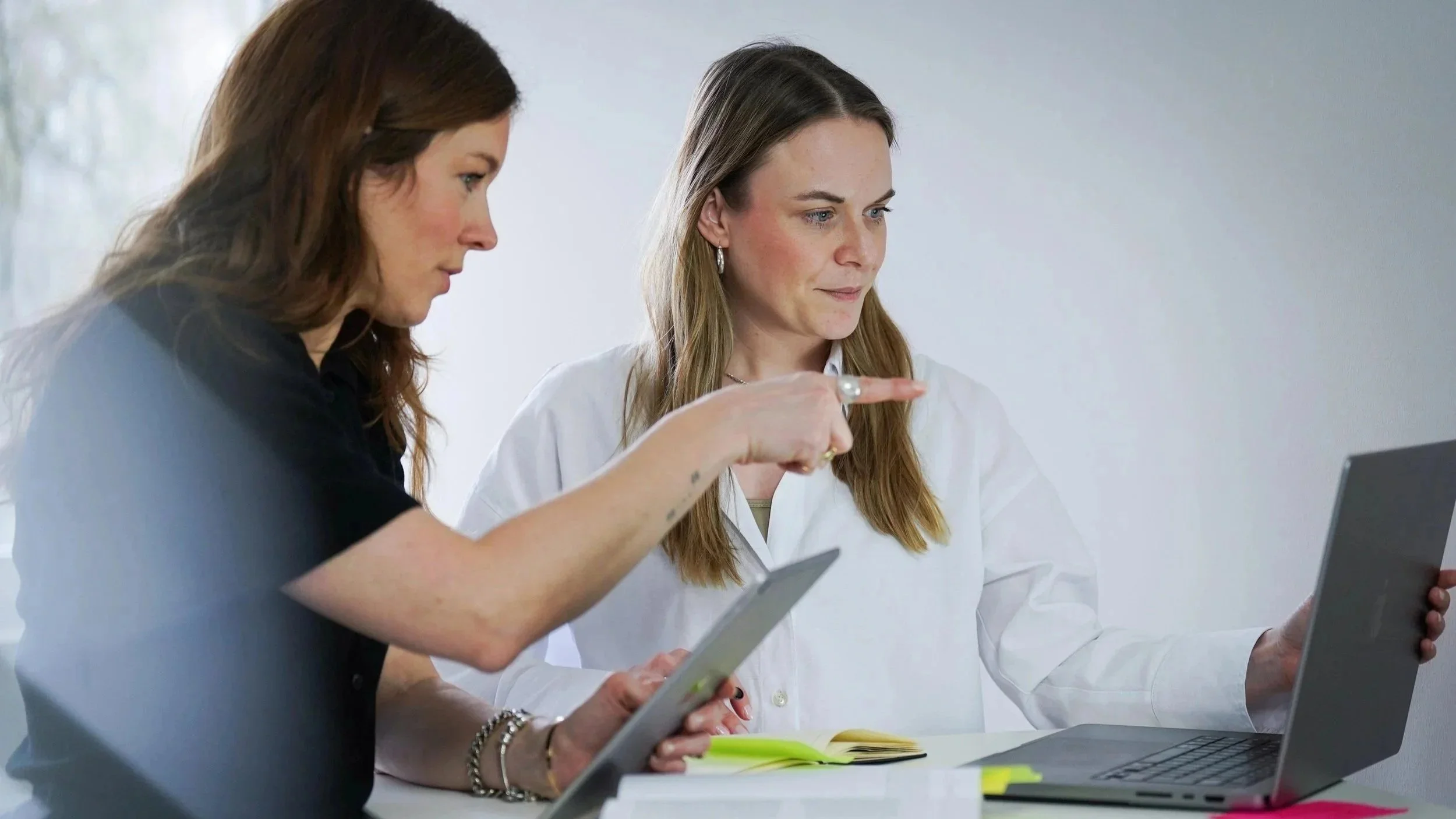 Zwei Frauen arbeiten an einem Tisch, eine mit Tablet, die andere mit Laptop, in einem grauen Raum.