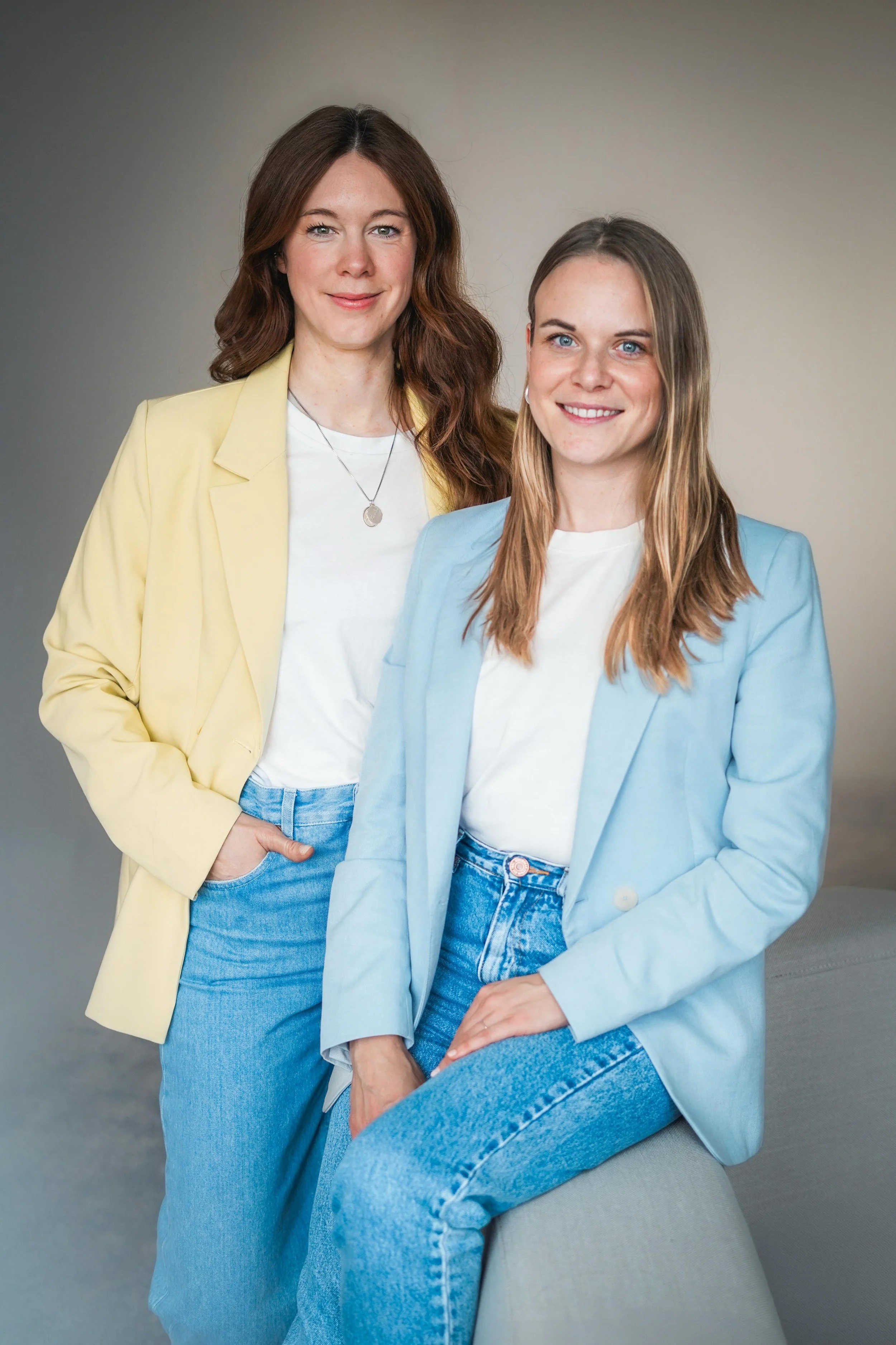 Zwei Frauen in pastelfarbenen Blazern, eine sitzt und die andere steht, vor grauem Hintergrund.