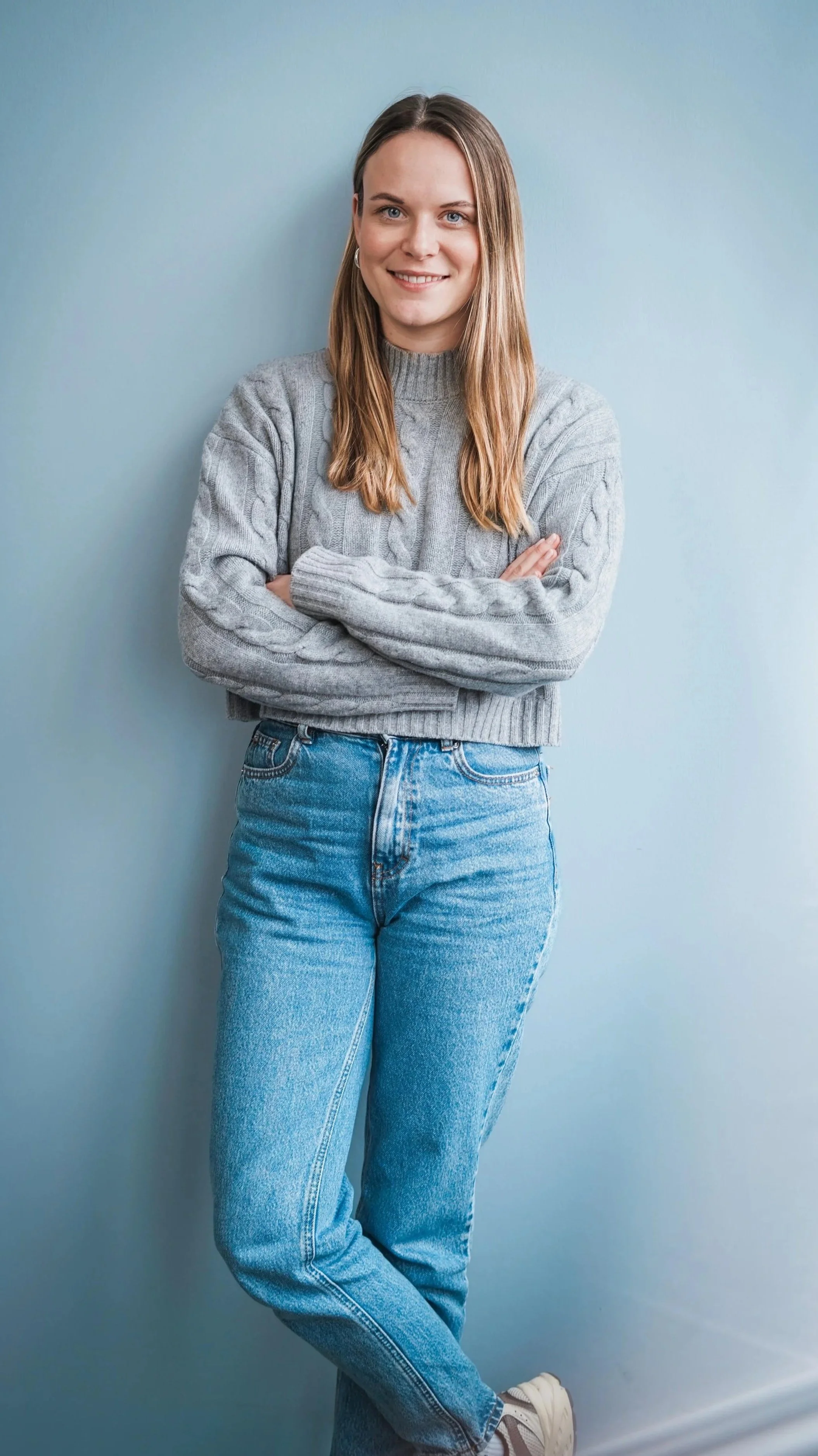 Junge Frau mit langem braunem Haar, grauem Pullover und blauen Jeans, steht mit verschränkten Armen vor einer hellblauen Wand.