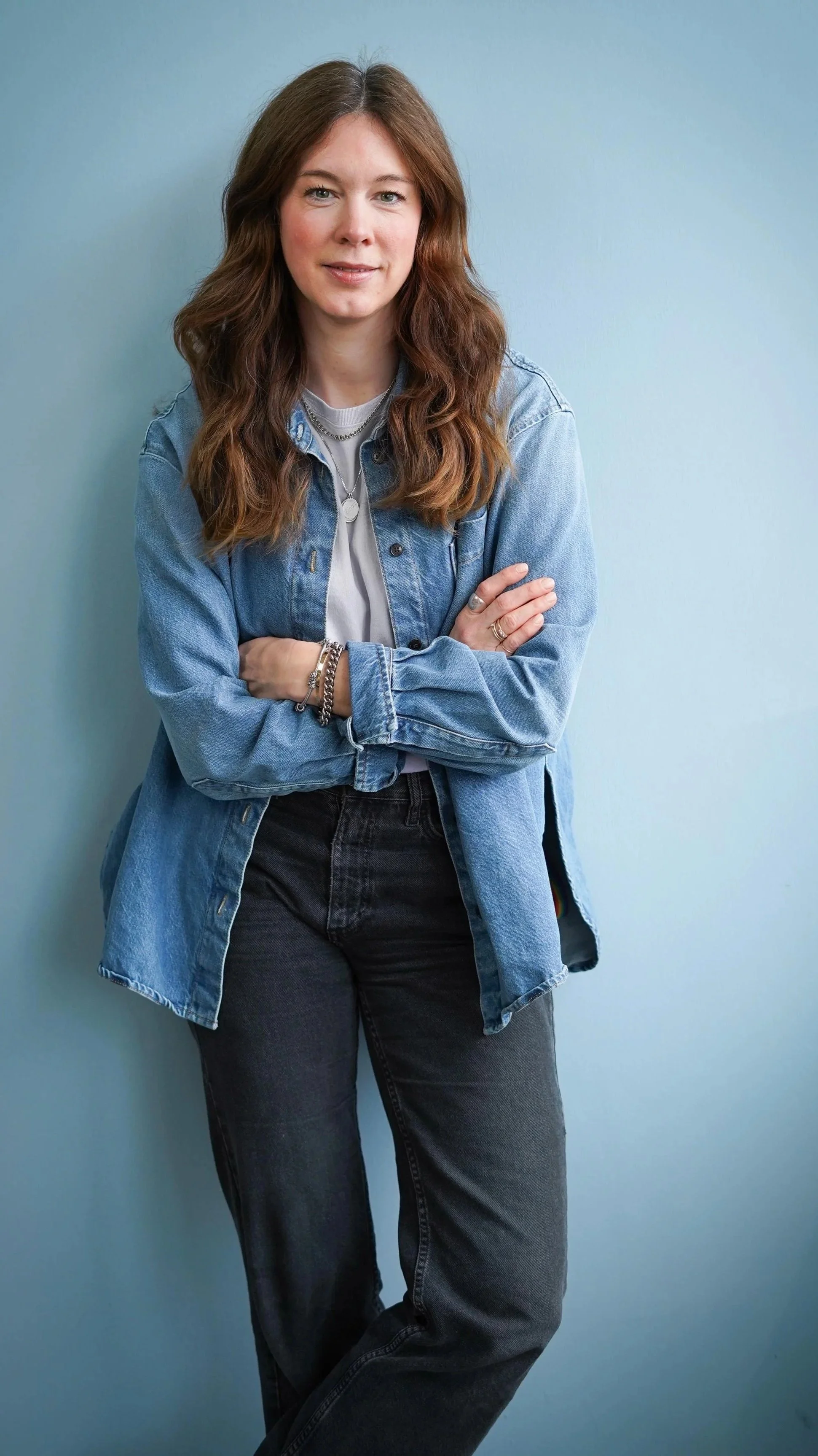 Junge Frau mit langen braunen Haaren, trägt eine Jeansjacke, T-Shirt und schwarze Jeans, steht vor einer hellblauen Wand und blickt lächelnd in die Kamera.