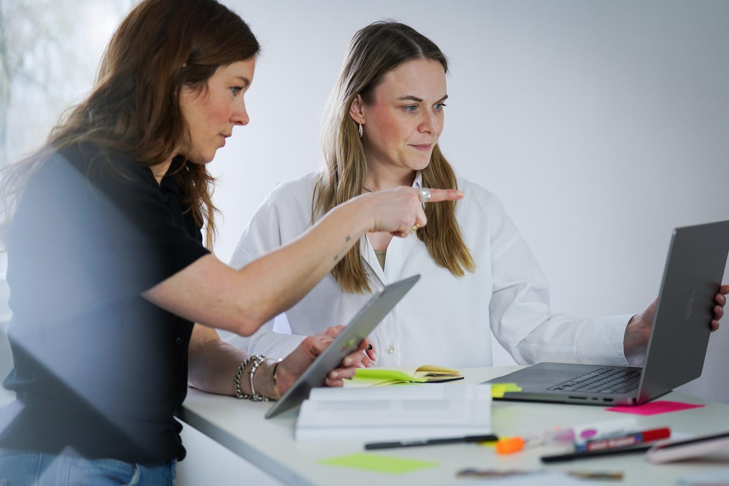 Zwei Frauen arbeiten gemeinsam an einem Laptop und einem Tablet in einem modernen Büro.