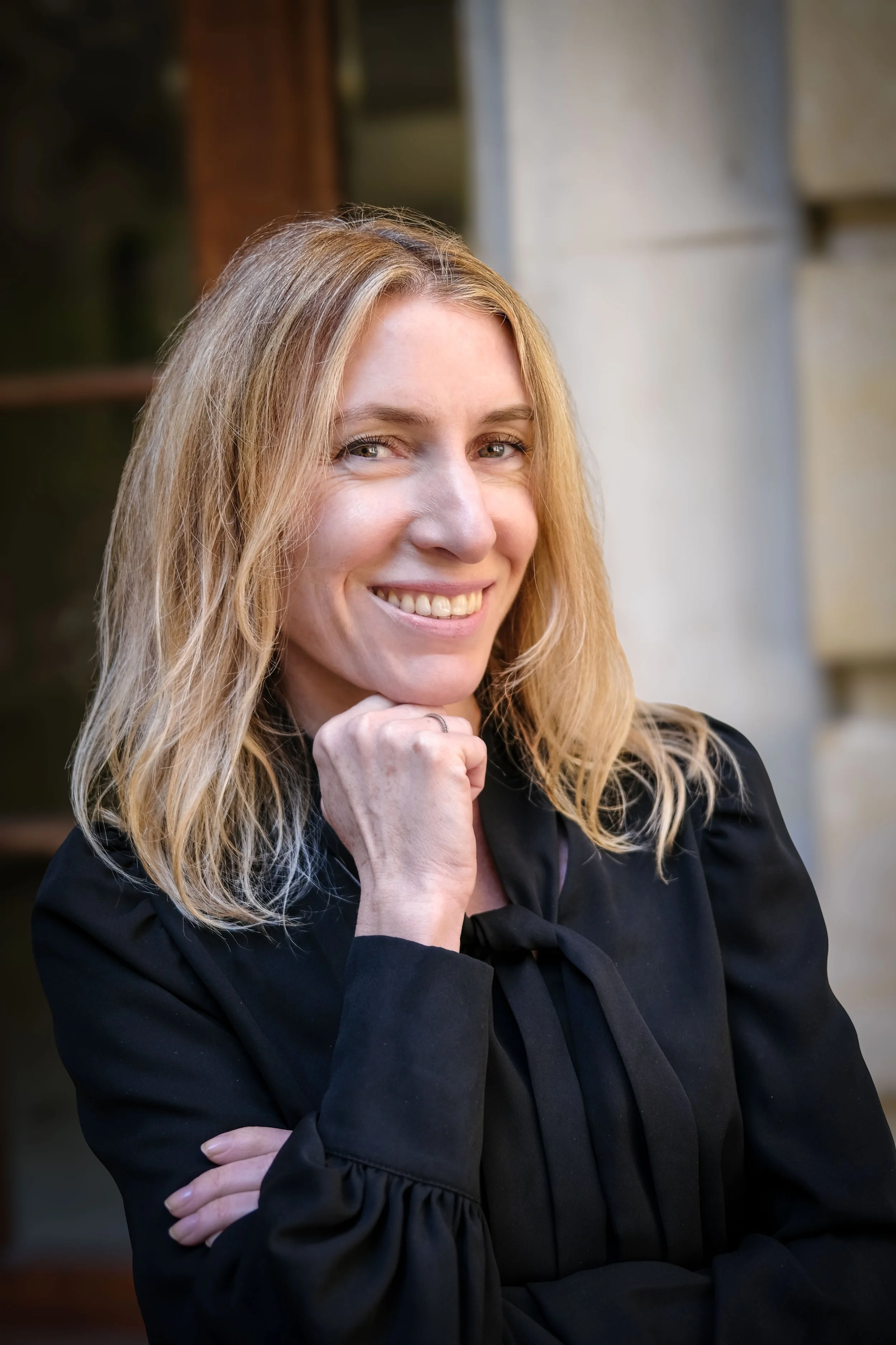 Une femme blonde souriante, portant une veste noire avec un nœud au col, pose en extérieur avec un fond flou