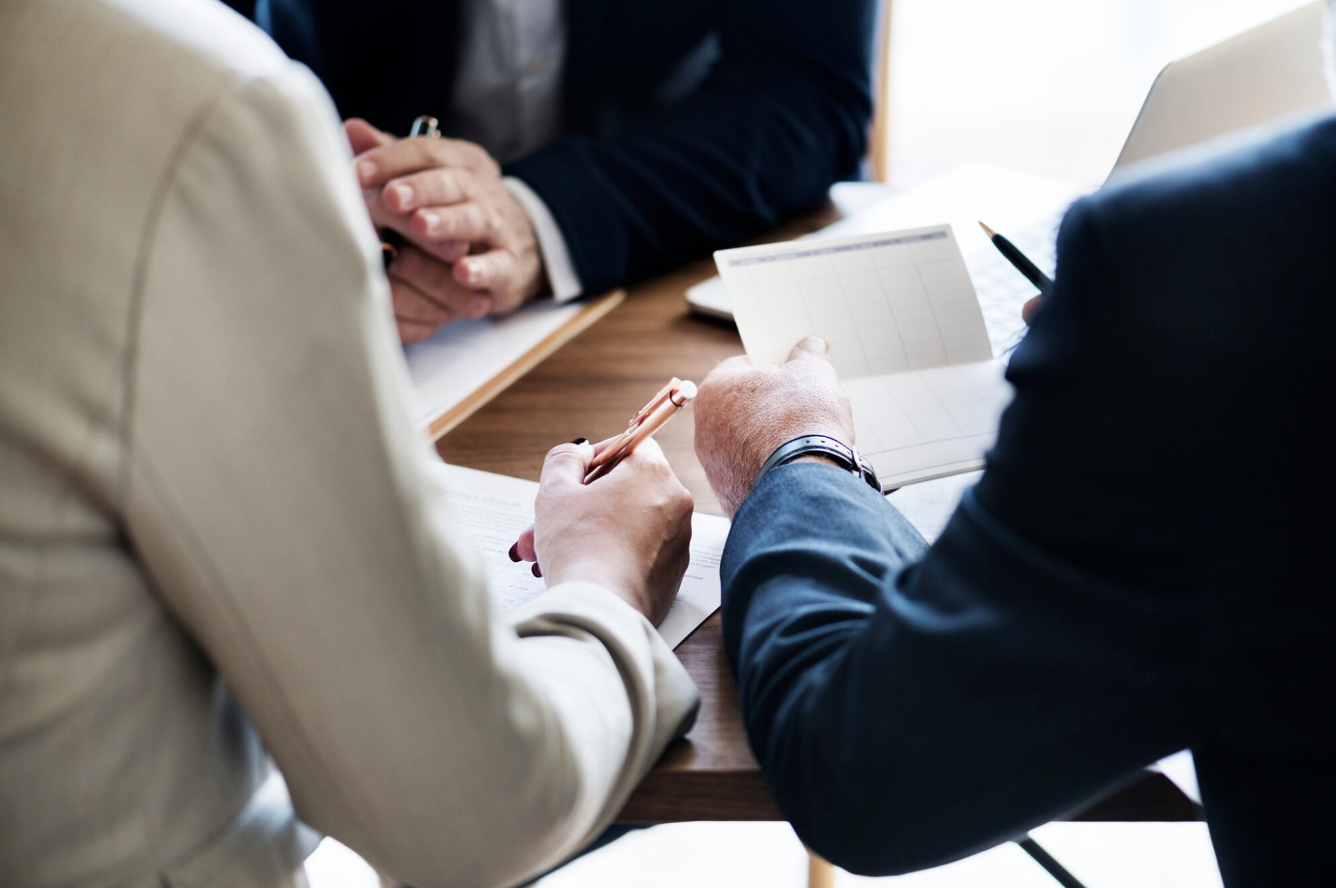 Trois personnes en costume discutant autour d'une table en bois, tenant des stylos et des papiers.