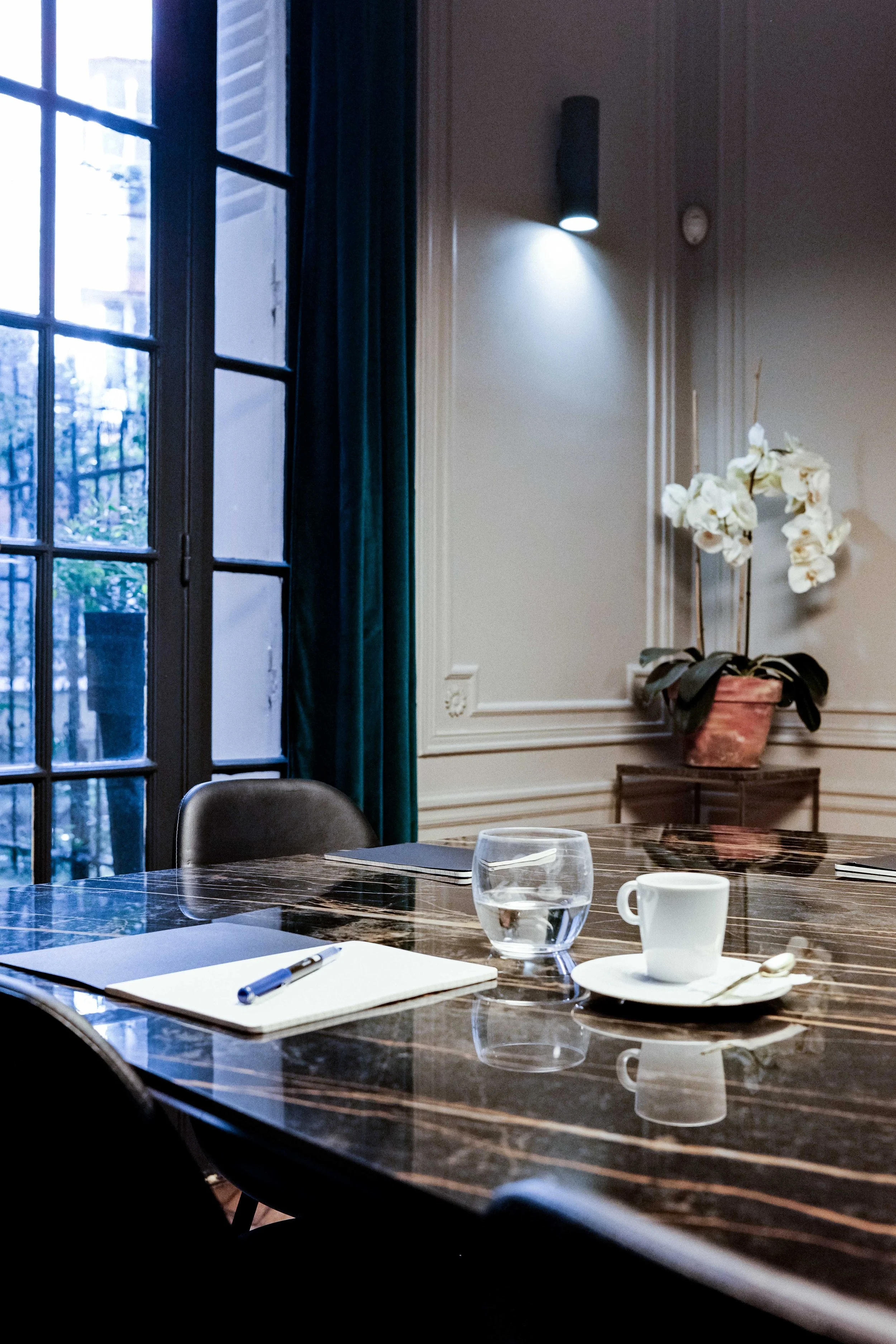 Une table en bois vitrée dressée dans une salle de réunion avec une tasse à café, un verre d'eau, un carnet et un stylo, une grande fenêtre avec rideaux bleus et une plante blanche dans un pot en terre cuite.