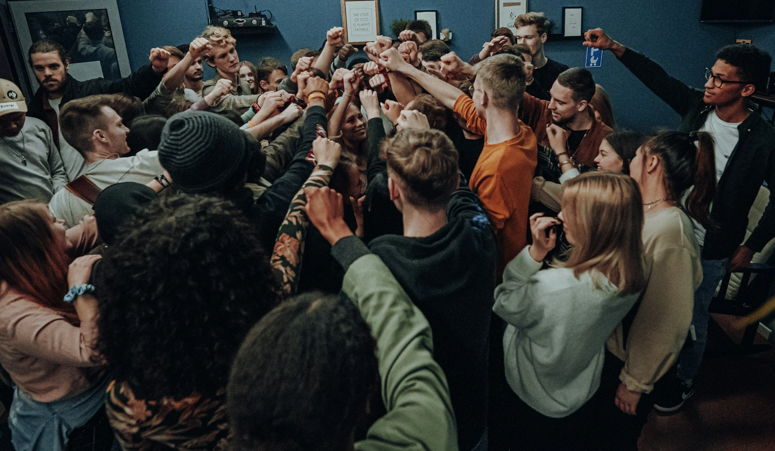 Group of young people gathered in a circle with raised fists, celebrating or supporting each other in a room with blue walls and framed pictures.